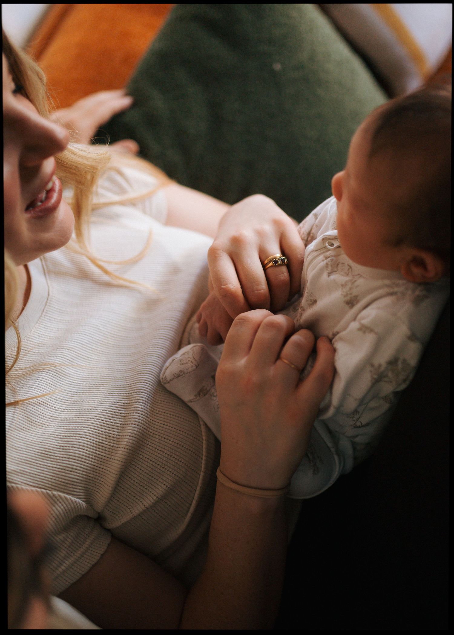 Close-up of a smiling mother with a gold ring gently holding the tiny hands of her newborn baby on a warm, cozy couch.