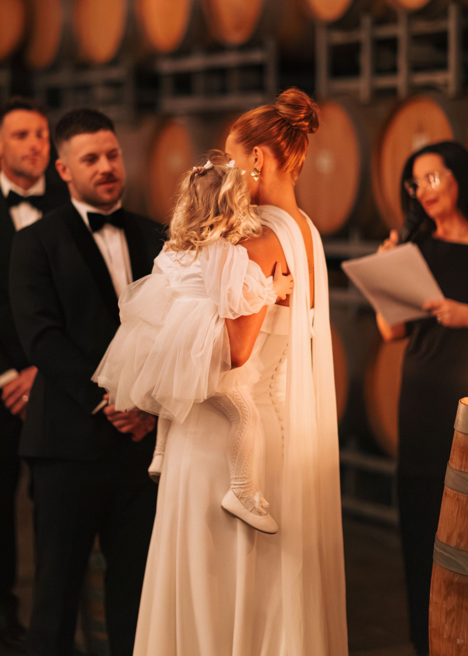 A bride in a white caped gown holds a young child during an indoor wedding ceremony surrounded by oak barrels.