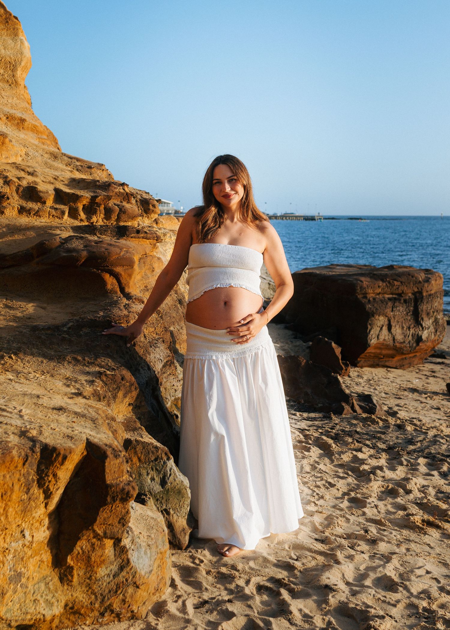 Smiling pregnant woman in white maternity outfit leans against golden sandstone rocks on sunny beach.