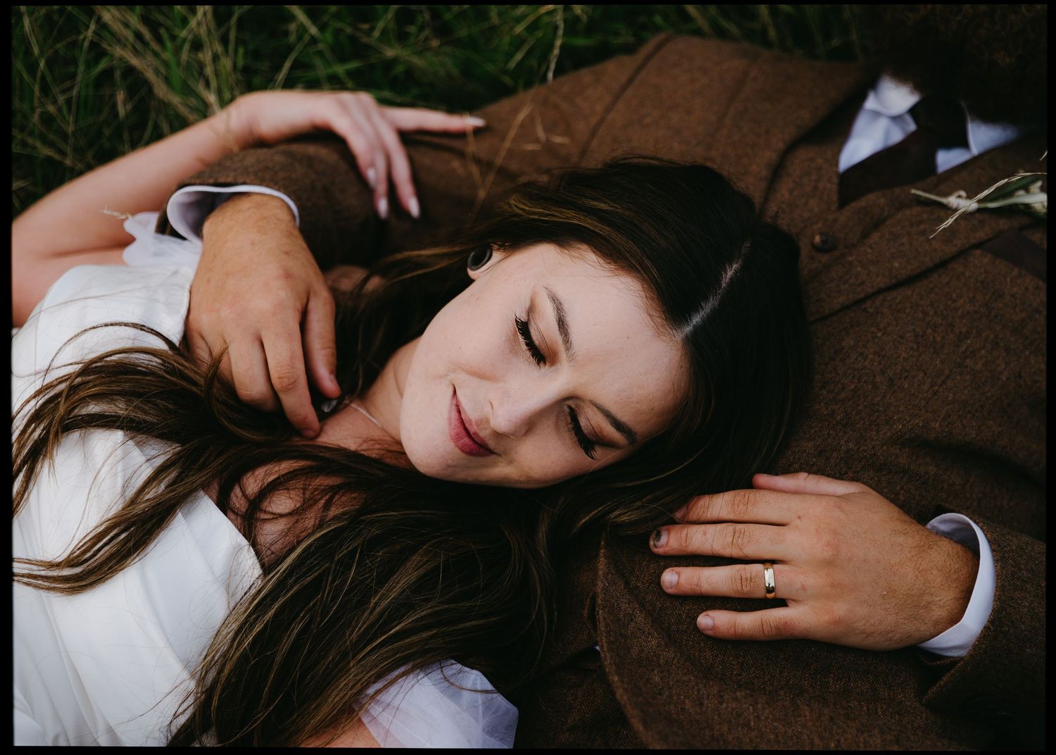 Bride resting her head on groom's chest while lying in green grass, wearing a gold wedding band.