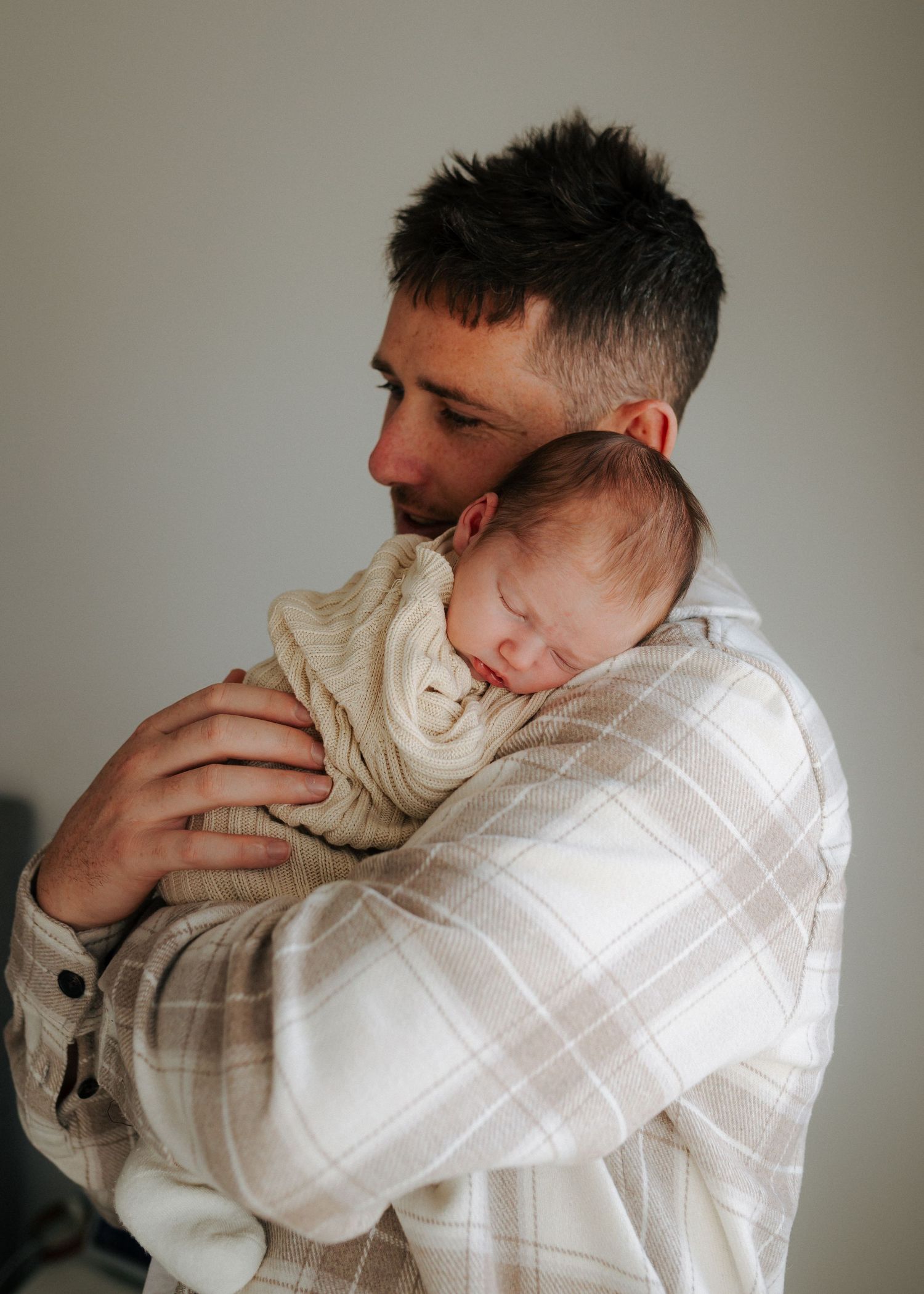 Father in plaid shirt gently holding sleeping newborn baby close to chest.