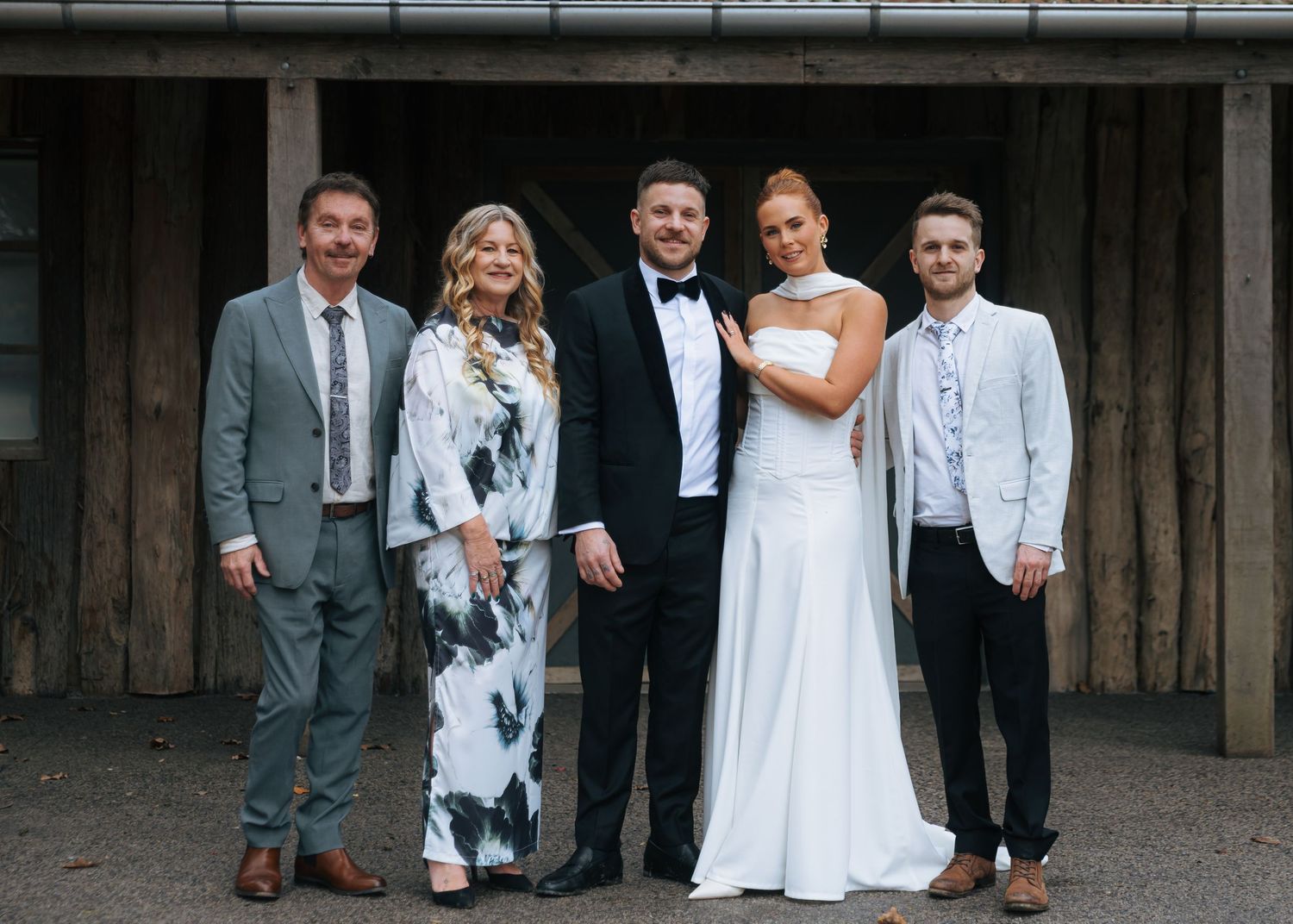 Wedding party of five posing together outside rustic venue, bride in white gown and groom in tuxedo.