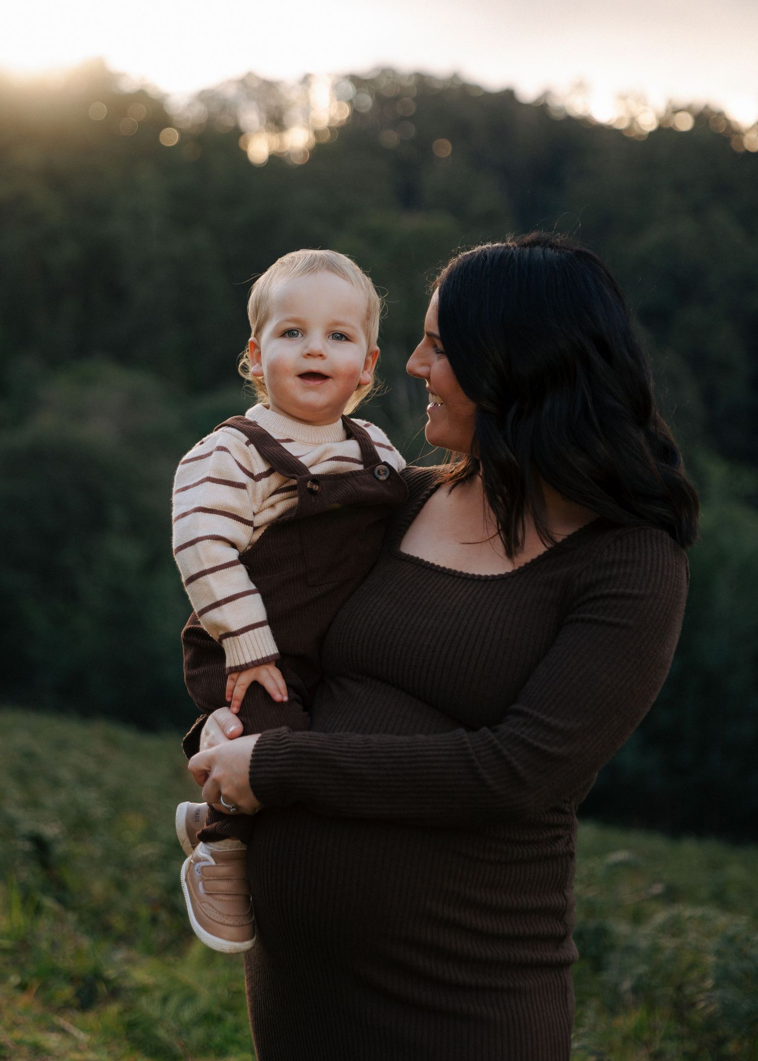 Pregnant mother in brown dress holding laughing toddler in green field during golden hour.
