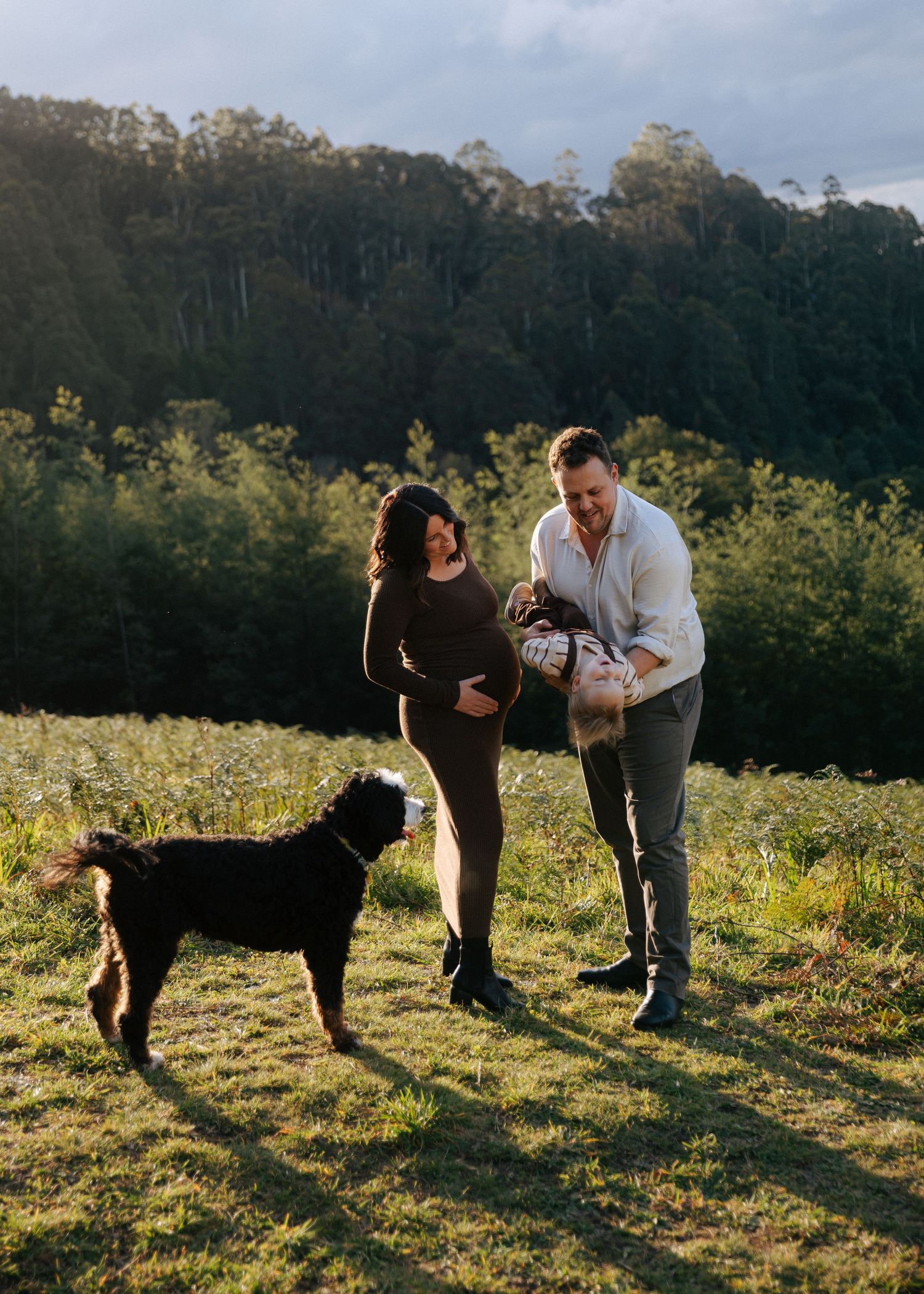 Couple with dog on sunny green hillside, man holding baby wrapped in blanket outdoors.