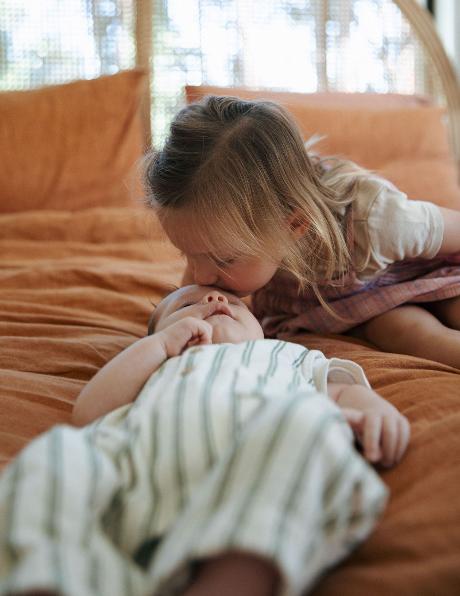 Young girl kissing her newborn baby sibling on an orange bed.