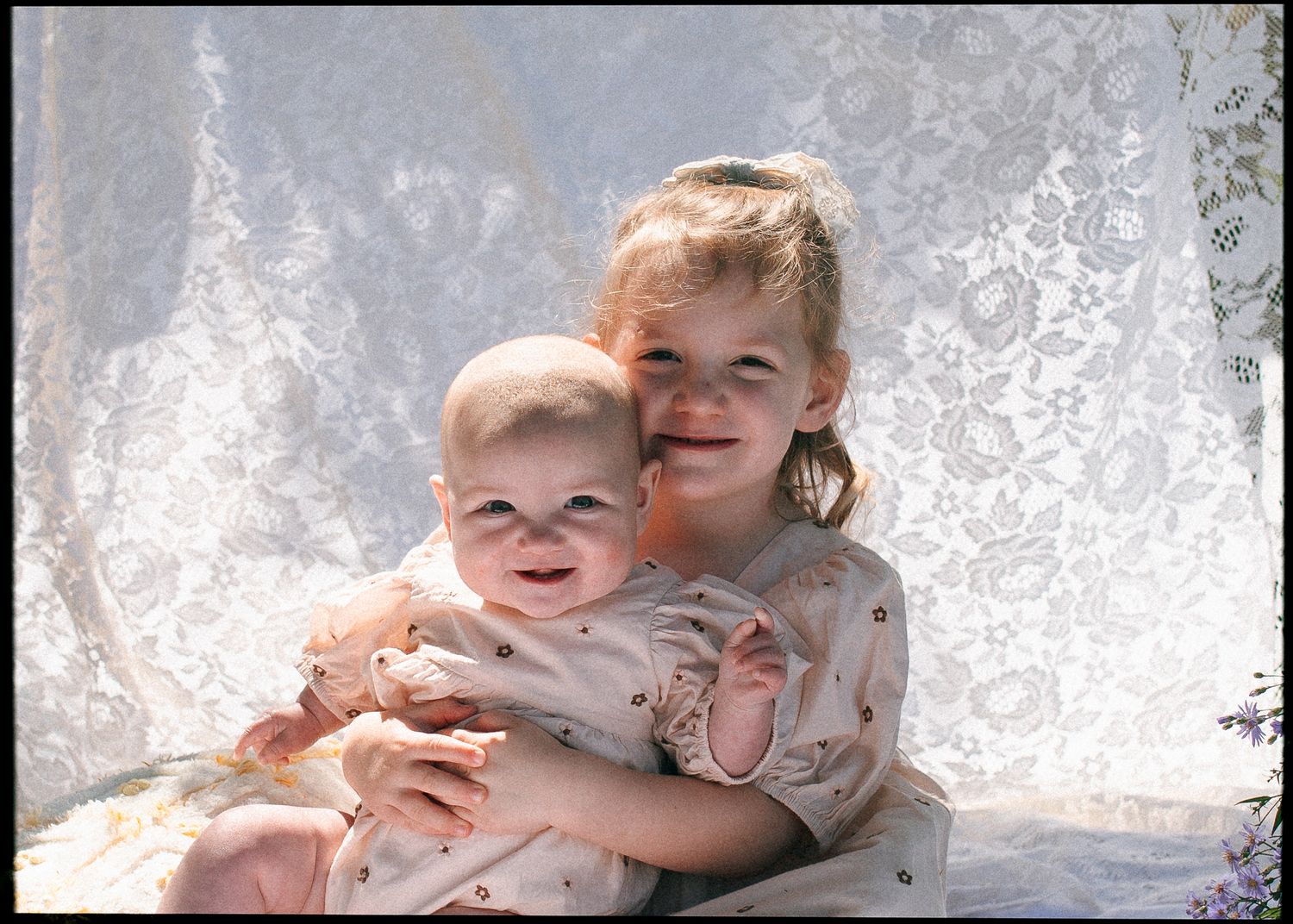Older sister hugging baby sibling in front of lace backdrop in vintage photo.