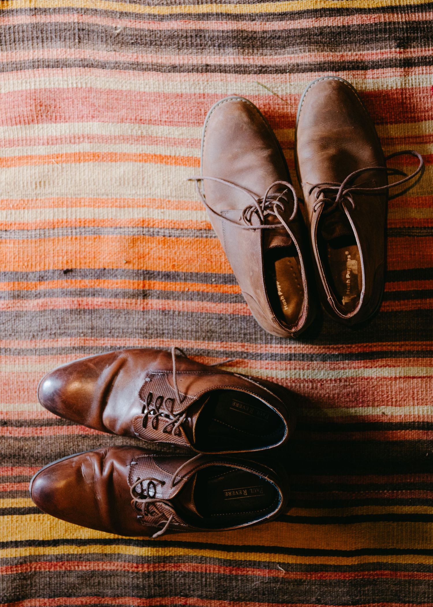 Two pairs of brown leather lace-up dress shoes on a colorful striped woven rug.