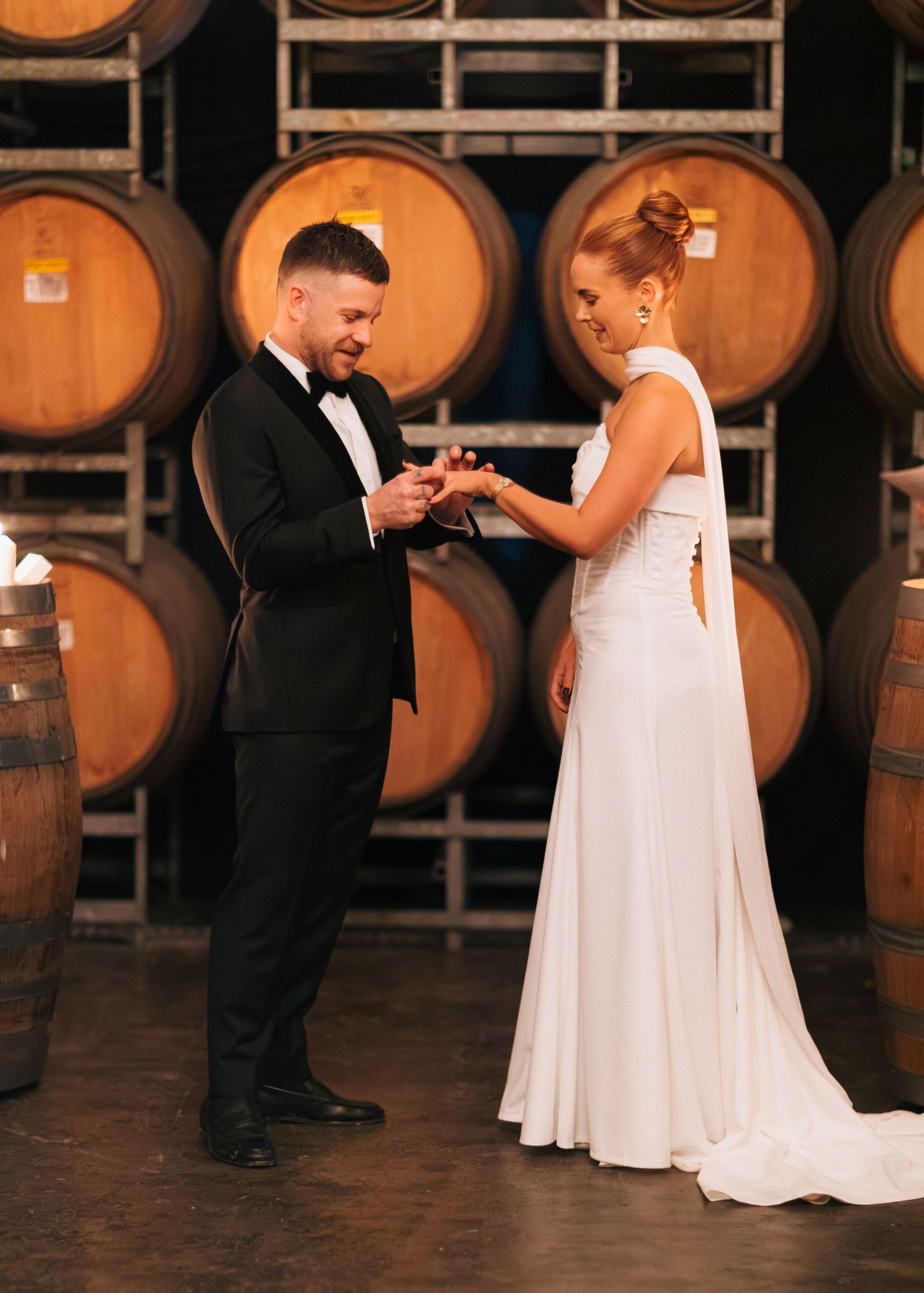 Groom places ring on brides finger during ceremony surrounded by oak wine barrels in winery.