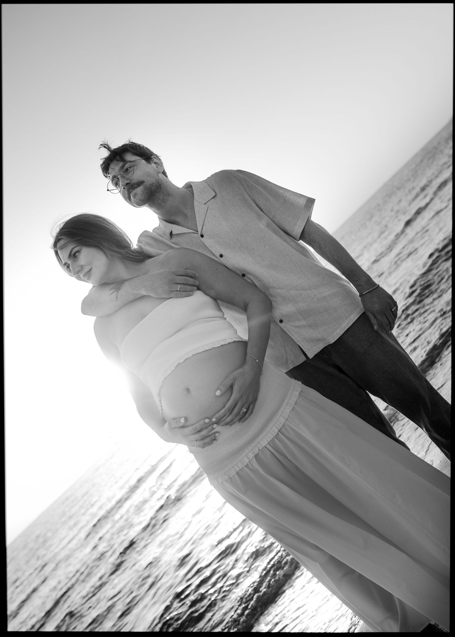 Black and white photo of a pregnant couple embracing by the ocean at sunset.