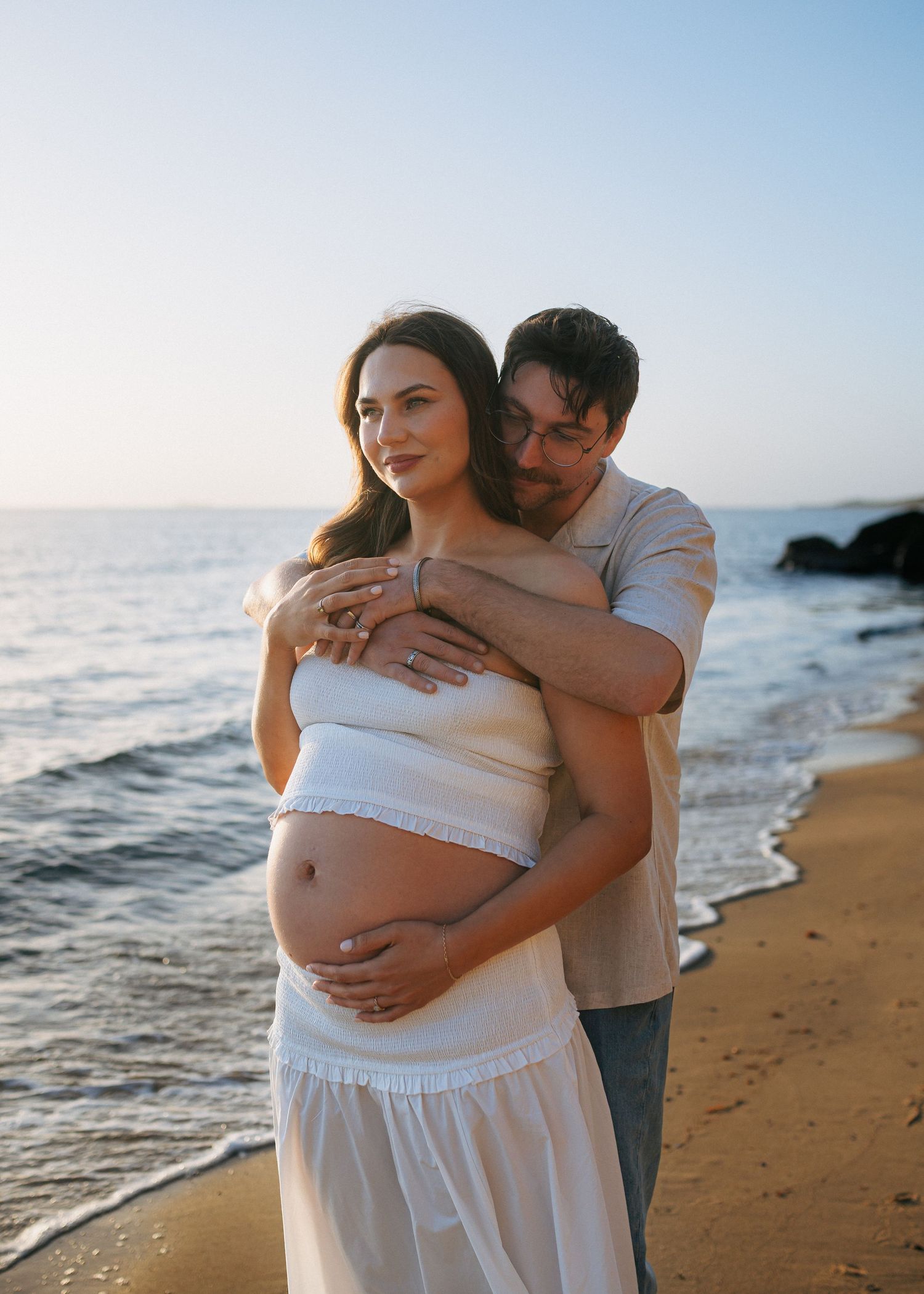 Couple embracing on beach at sunset, man holding pregnant womans baby bump near ocean waves.
