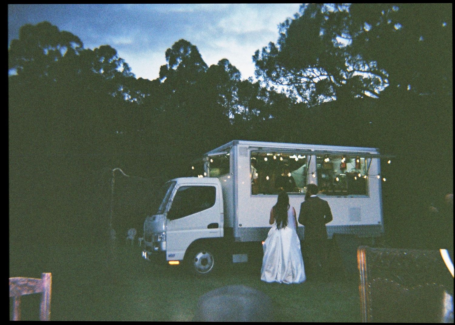 Bride and groom stand before an illuminated food truck at dusk surrounded by tall trees at their wedding.