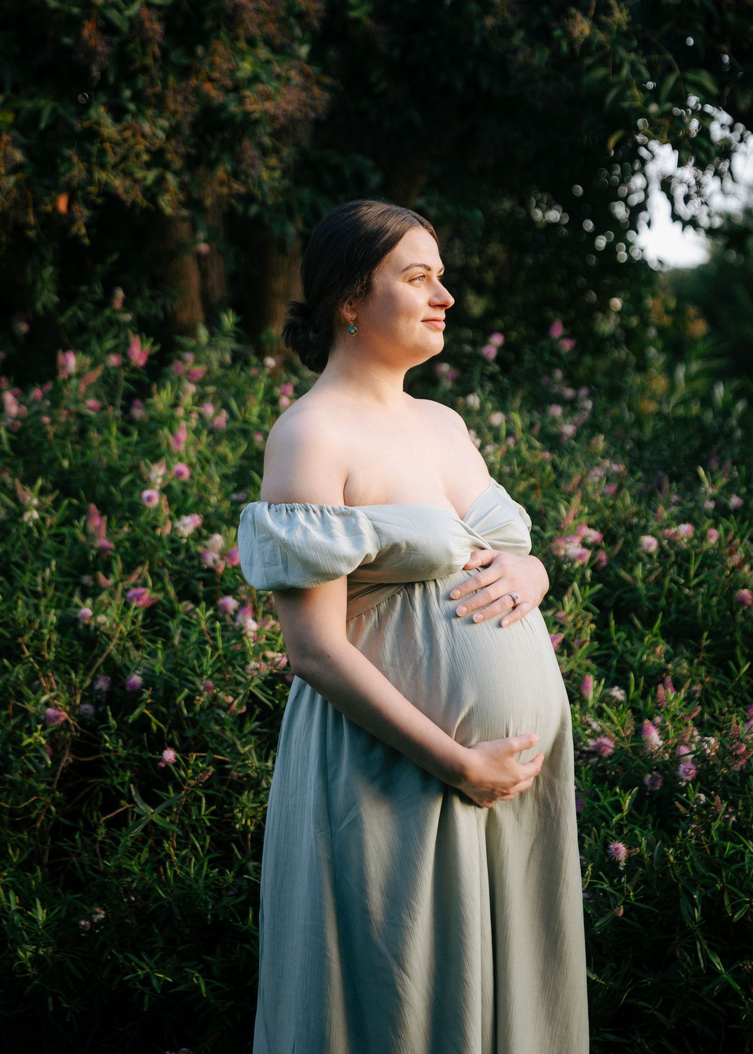 Pregnant woman in sage off-shoulder gown holding baby bump among pink flowering bushes in golden hour light.