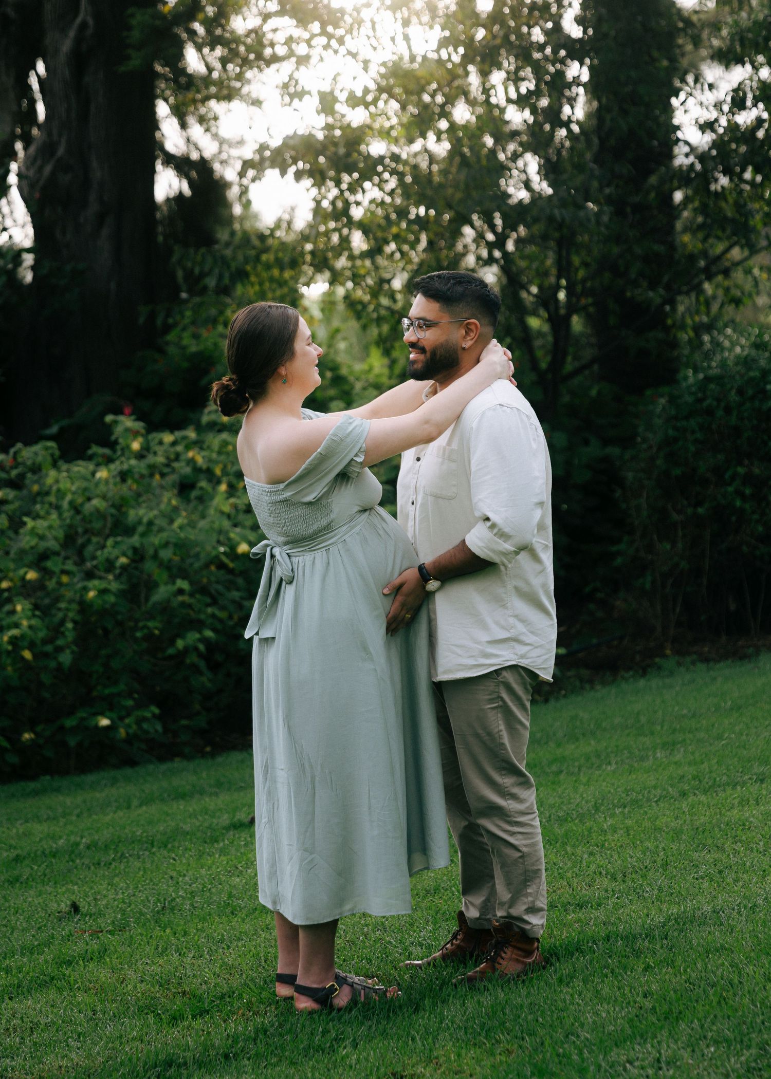 Expecting couple sharing a tender moment on green lawn, woman in sage dress touching partner's face.