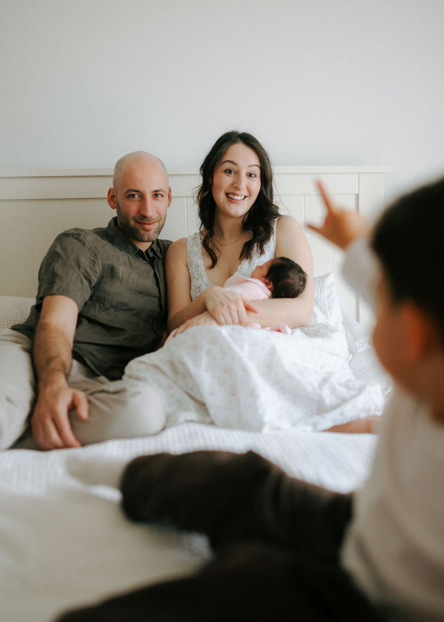 Smiling parents on bed with newborn as toddler gestures in foreground during family session.
