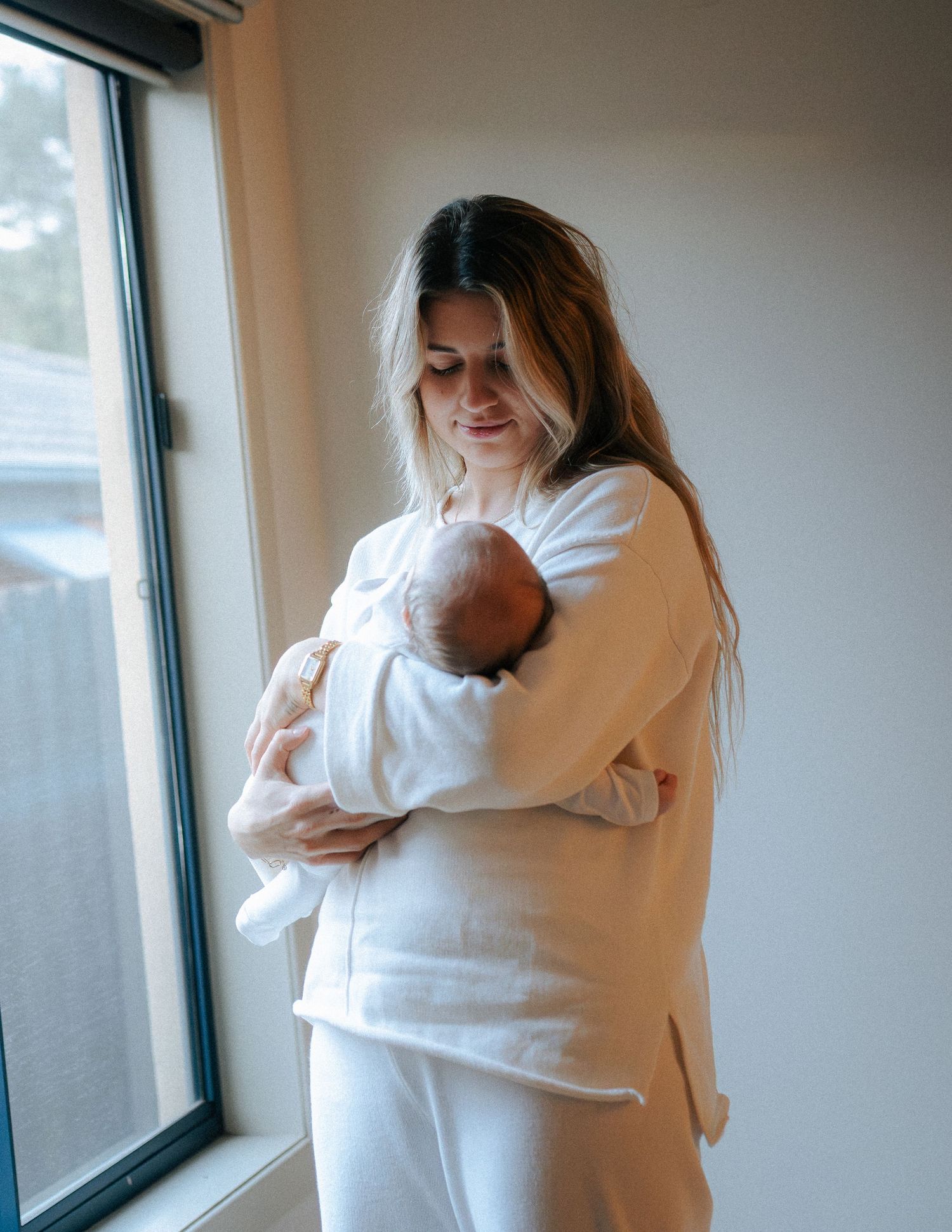 New mother in white outfit smiling while gently holding sleeping newborn by window.
