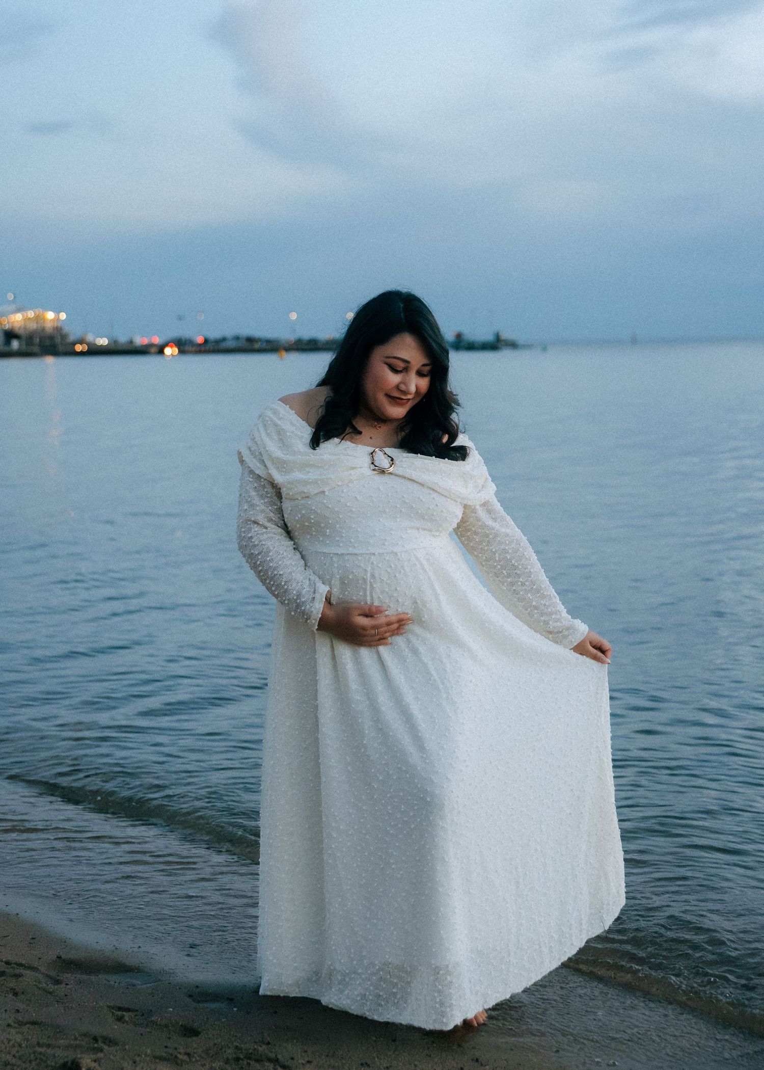 Pregnant woman in white lace maxi dress cradling baby bump while standing at water's edge.