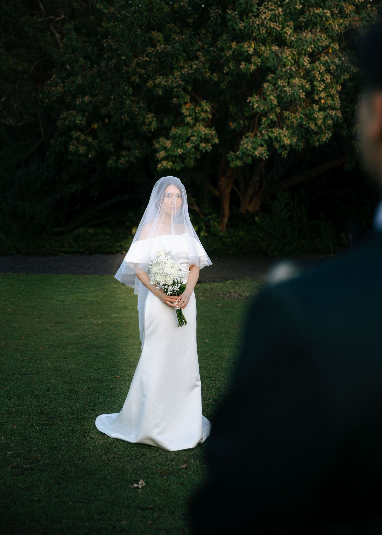 Elegant bride in off-shoulder white gown and veil holding white bouquet standing on lush green lawn at dusk.