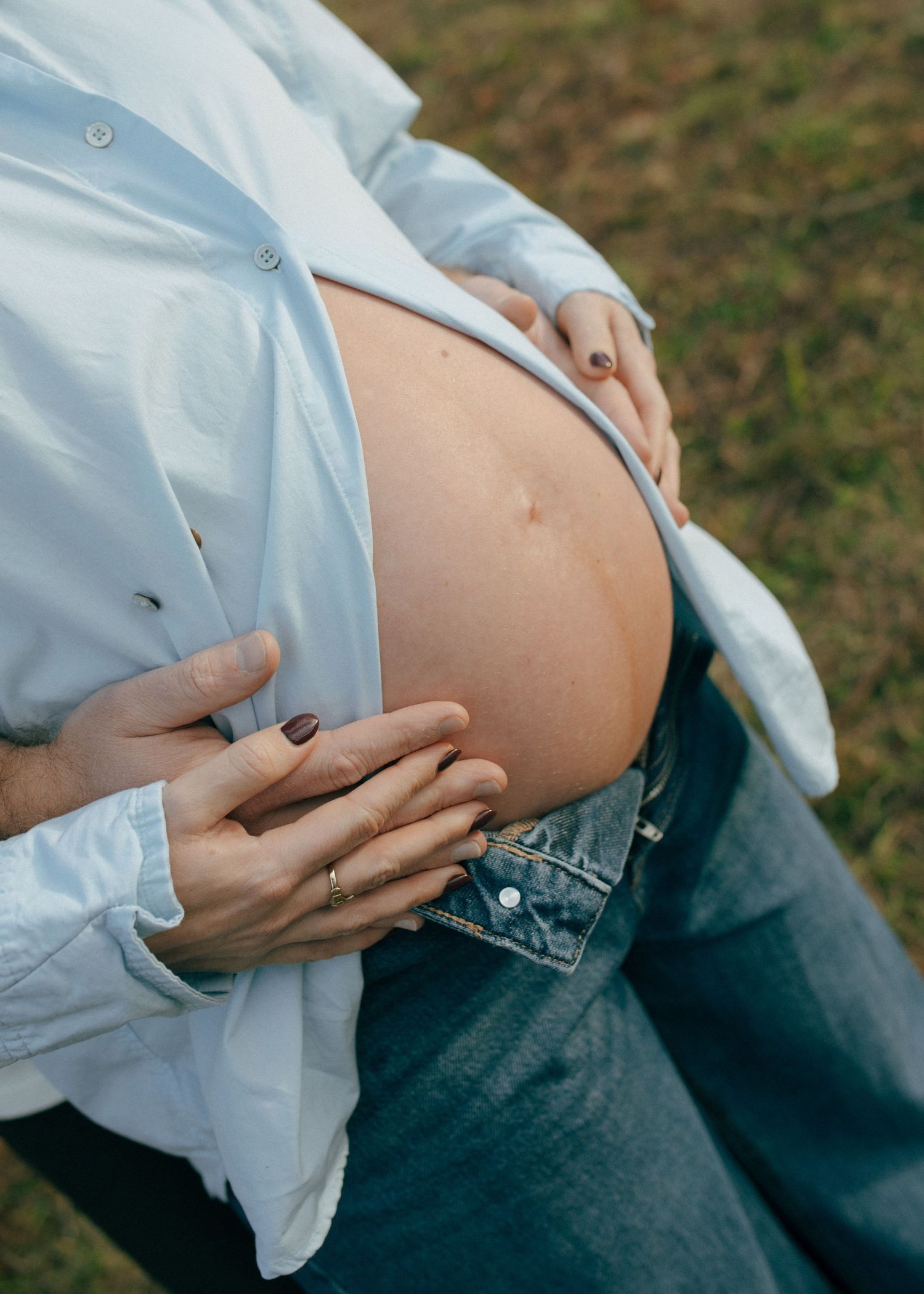 Detail shot of pregnant womans bare baby bump with hands resting on belly during outdoor maternity session.