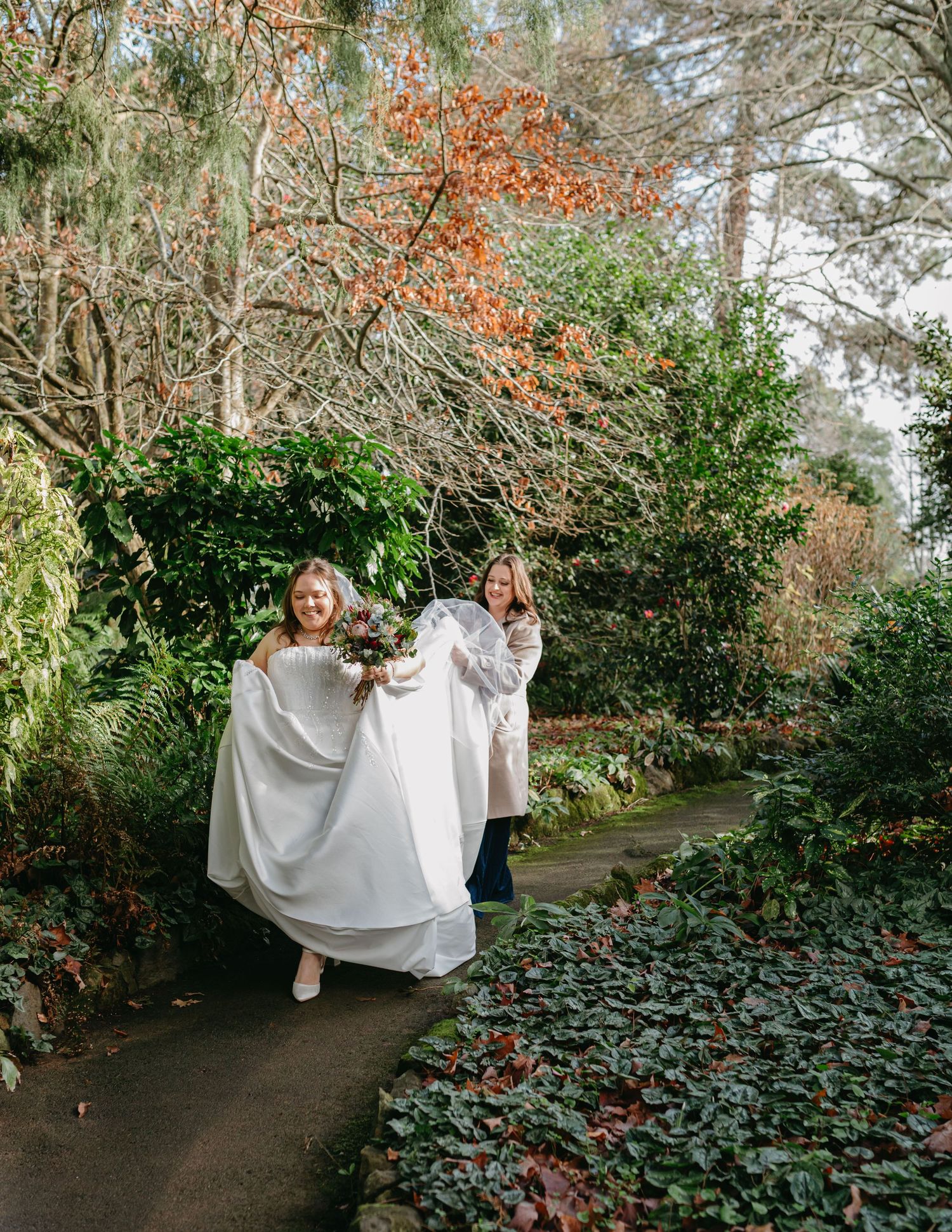 Bride in white gown walking garden path with assistant carrying train among autumn foliage.