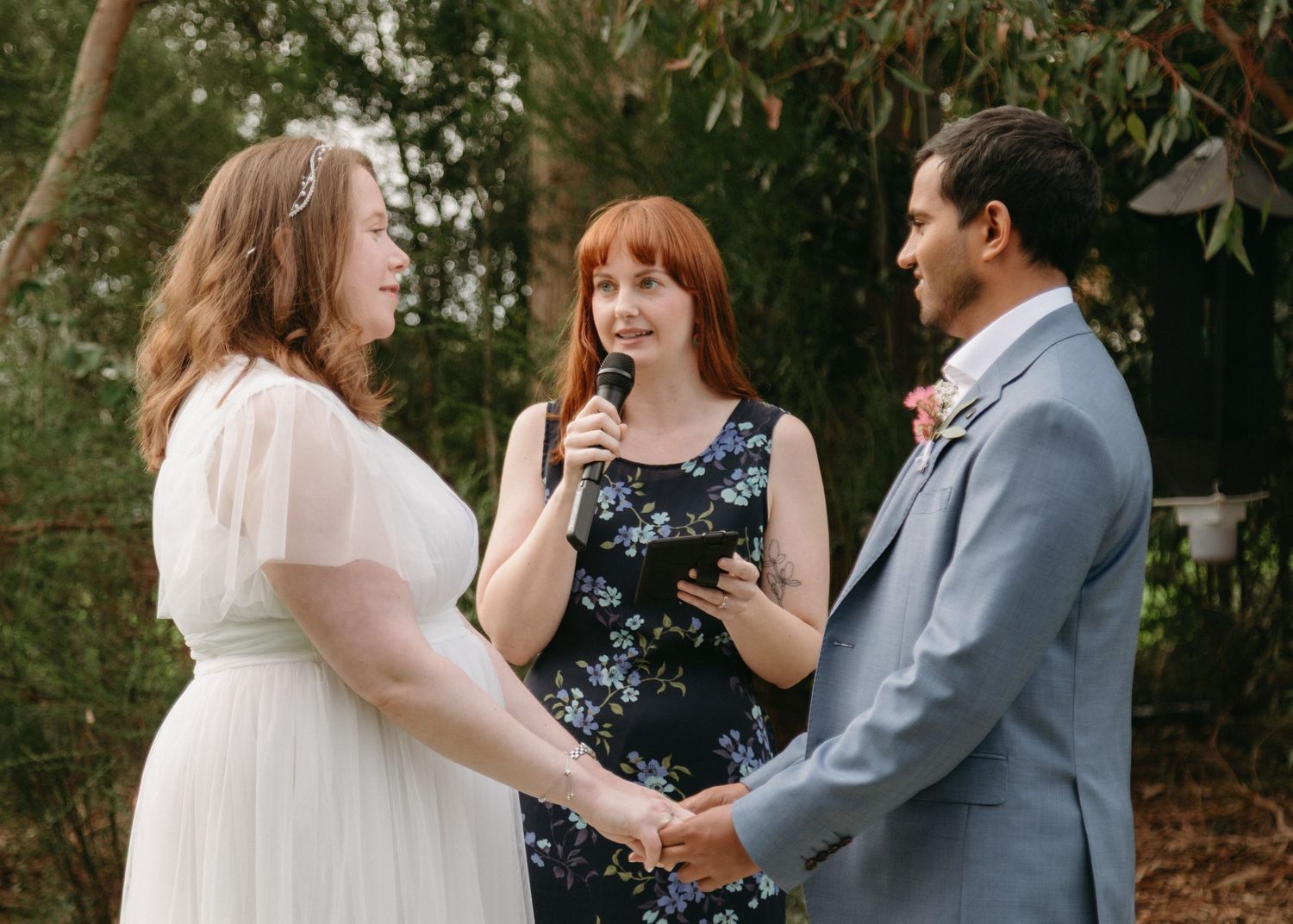Wedding officiant reads vows as bride and groom hold hands at outdoor garden ceremony.
