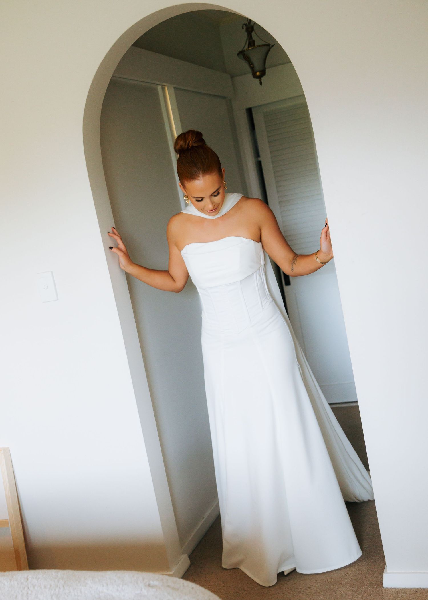 Bride in white strapless gown with train stands in arched doorway looking down.