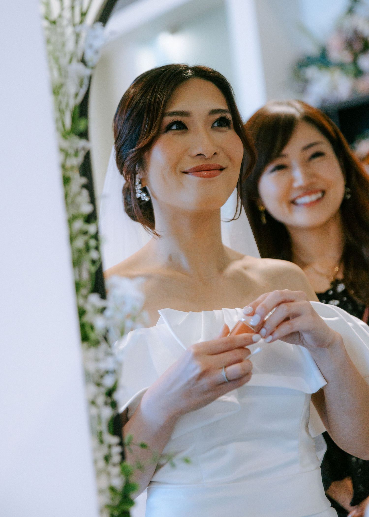 Elegant bride in off-shoulder white dress with updo hairstyle and drop earrings smiling at ceremony.
