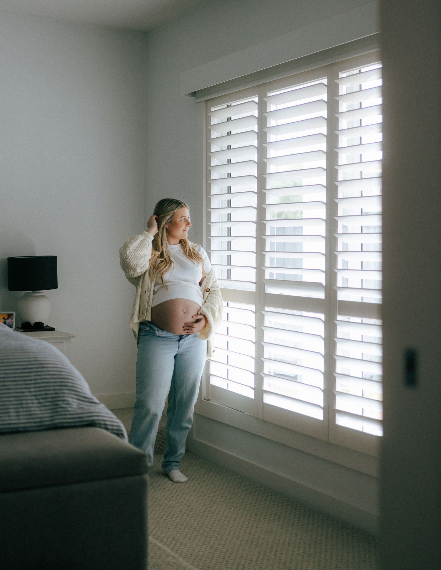 Pregnant woman in white top and jeans standing by bright plantation shutters holding her baby bump.