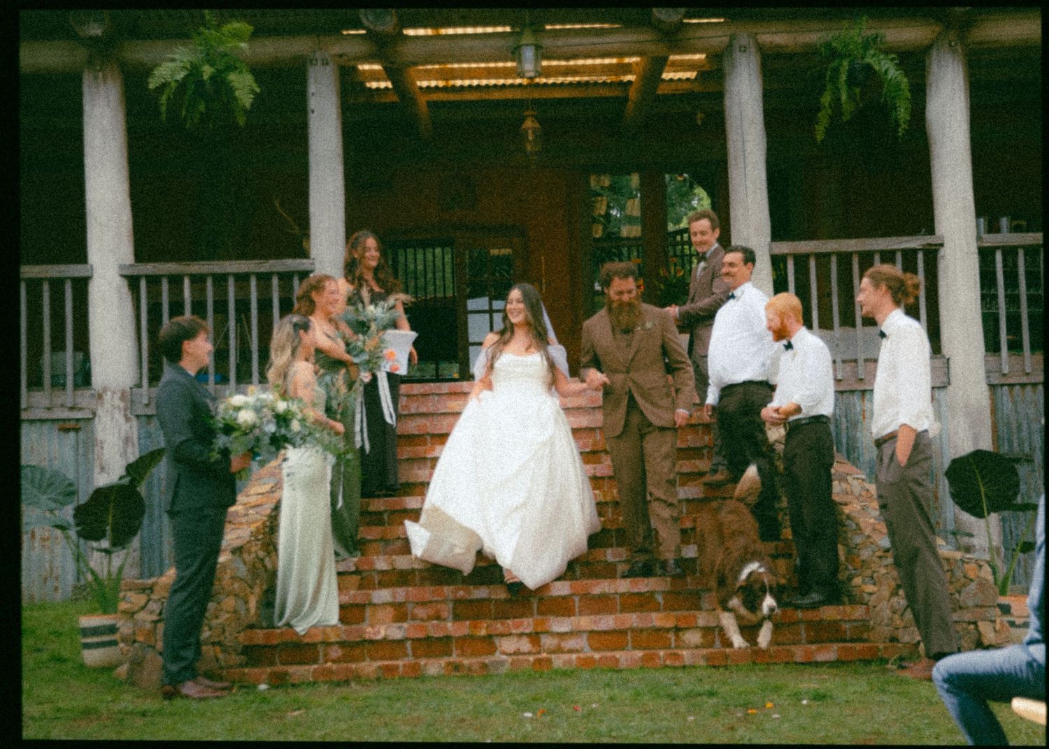 Bride in white gown descends brick steps with wedding party at a rustic outdoor venue.