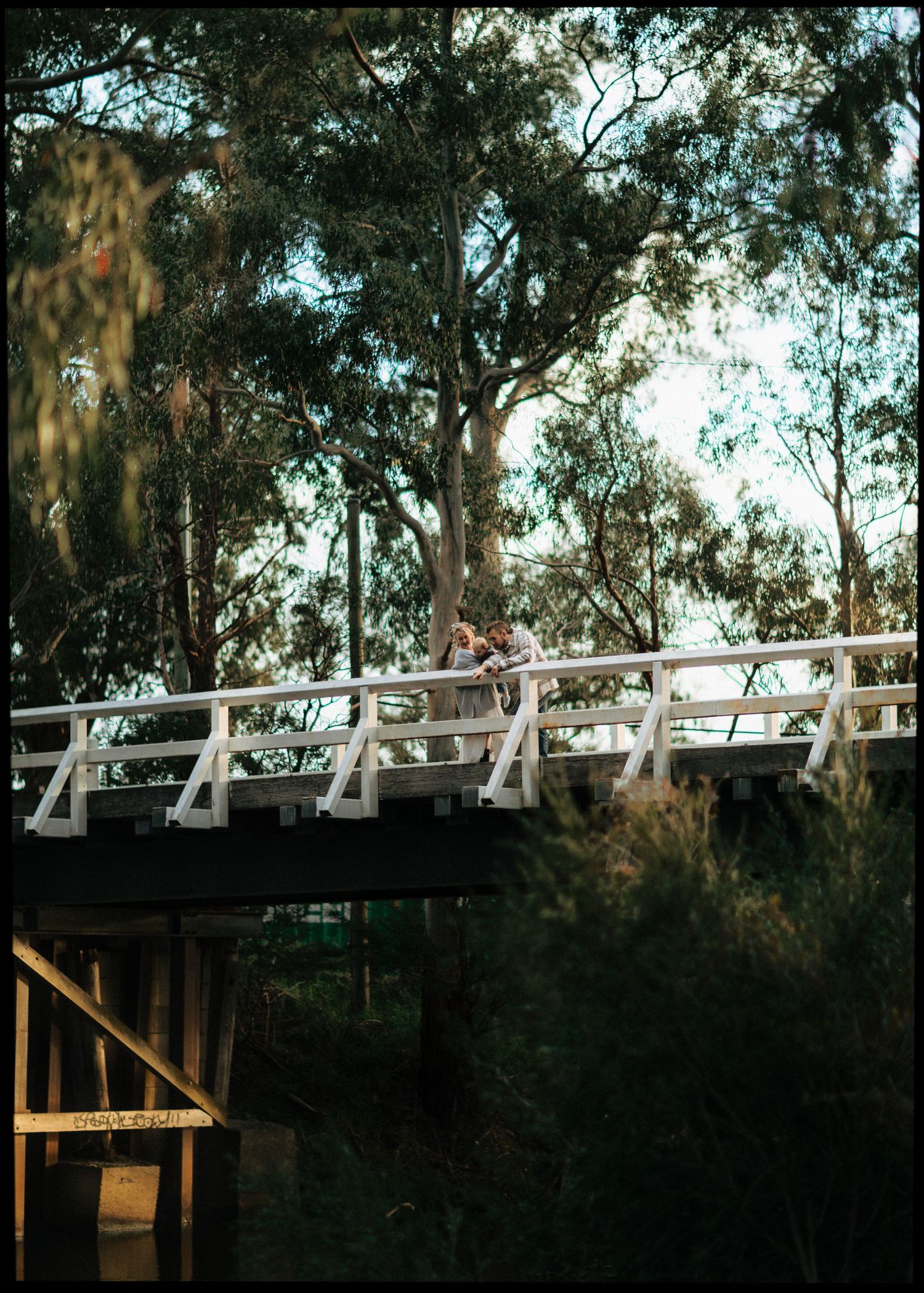 Family standing together on a white wooden bridge surrounded by lush eucalyptus trees.