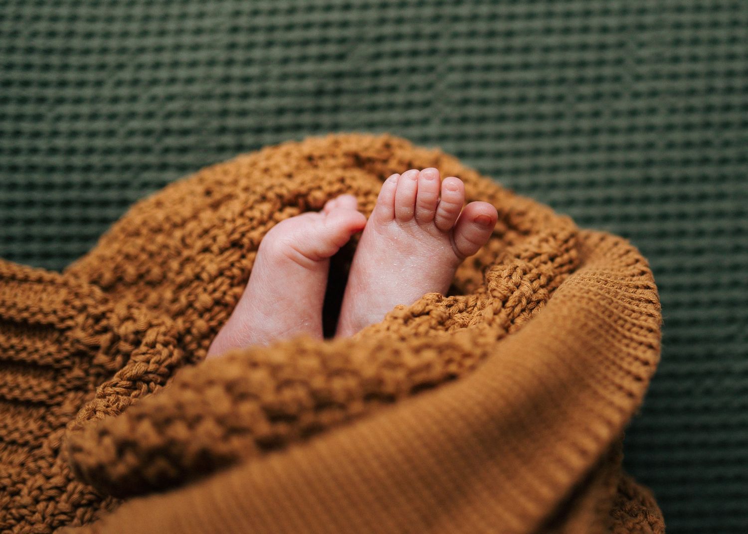 Tiny newborn baby feet peeking out from a cozy mustard knit blanket on green fabric.