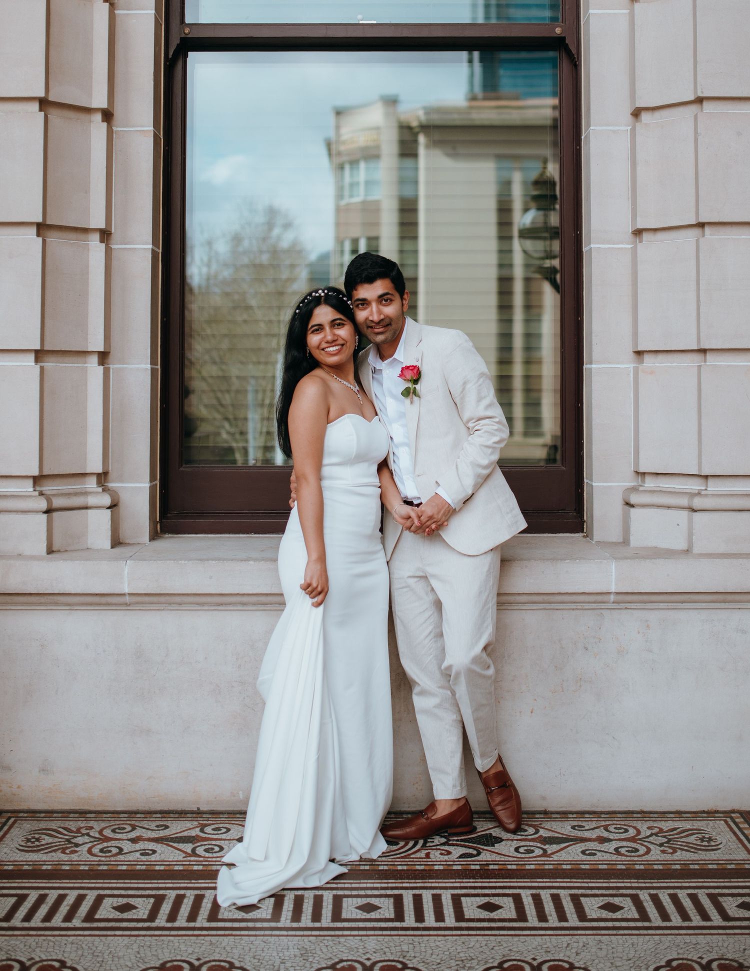 Elegant bride and groom portrait in white wedding attire posing before ornate building columns.