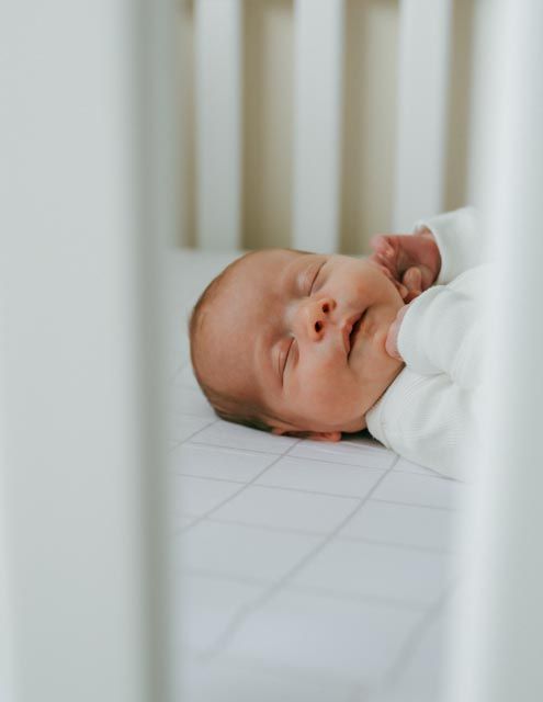 Sleeping newborn baby in white onesie lying in a white crib.