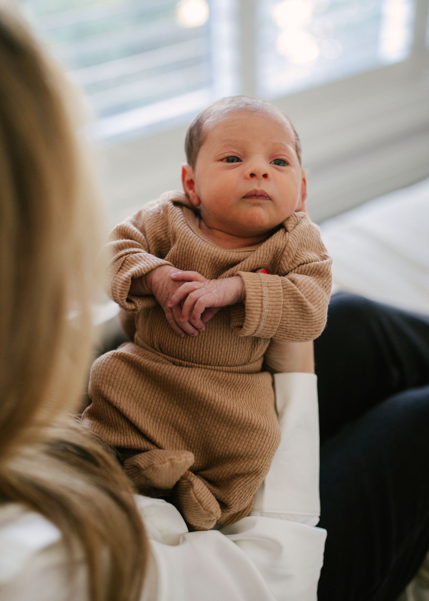 Alert newborn baby in brown ribbed romper sitting up while being held by mother.