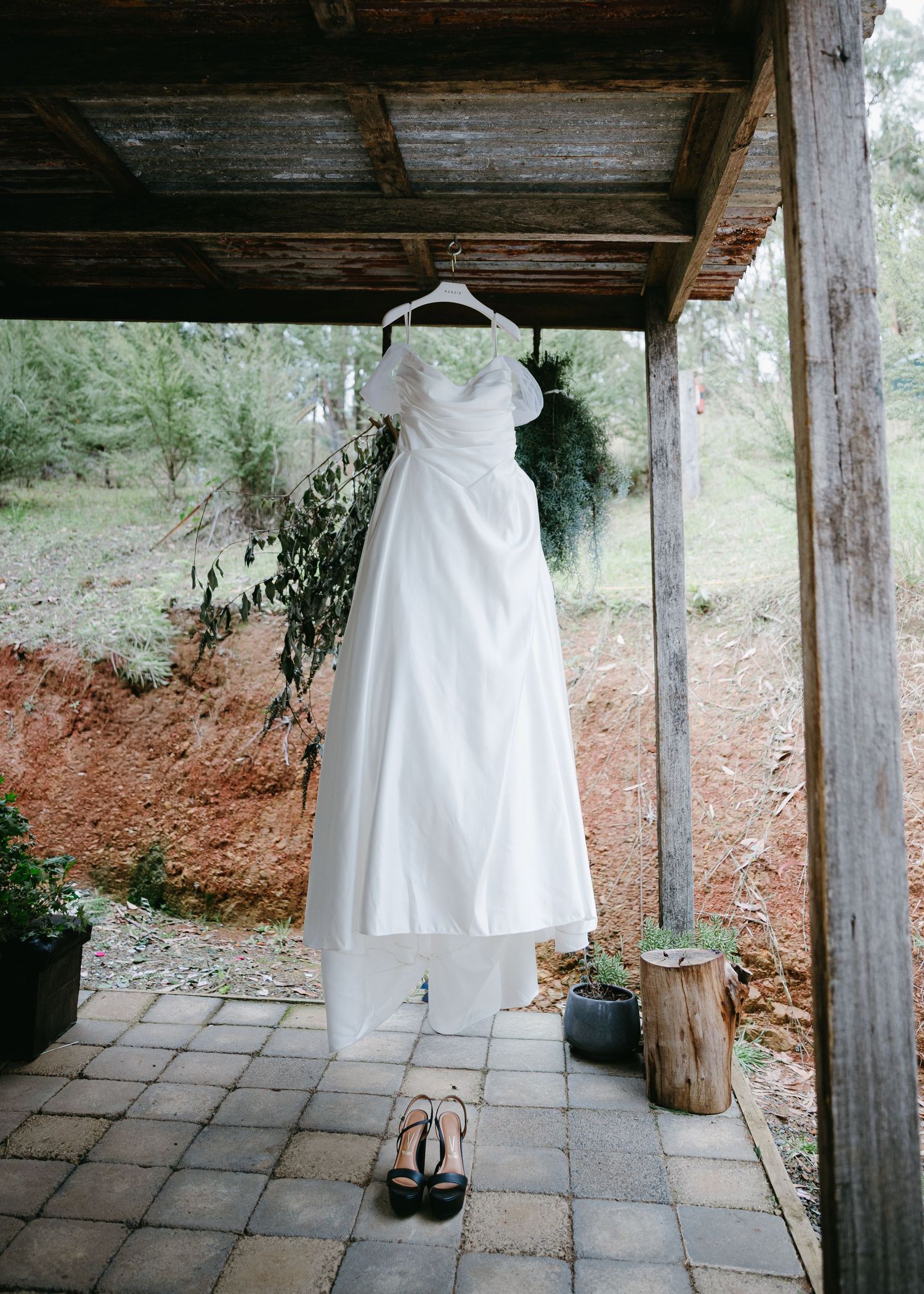 Elegant white wedding dress hanging on rustic wooden porch with black heels placed below.