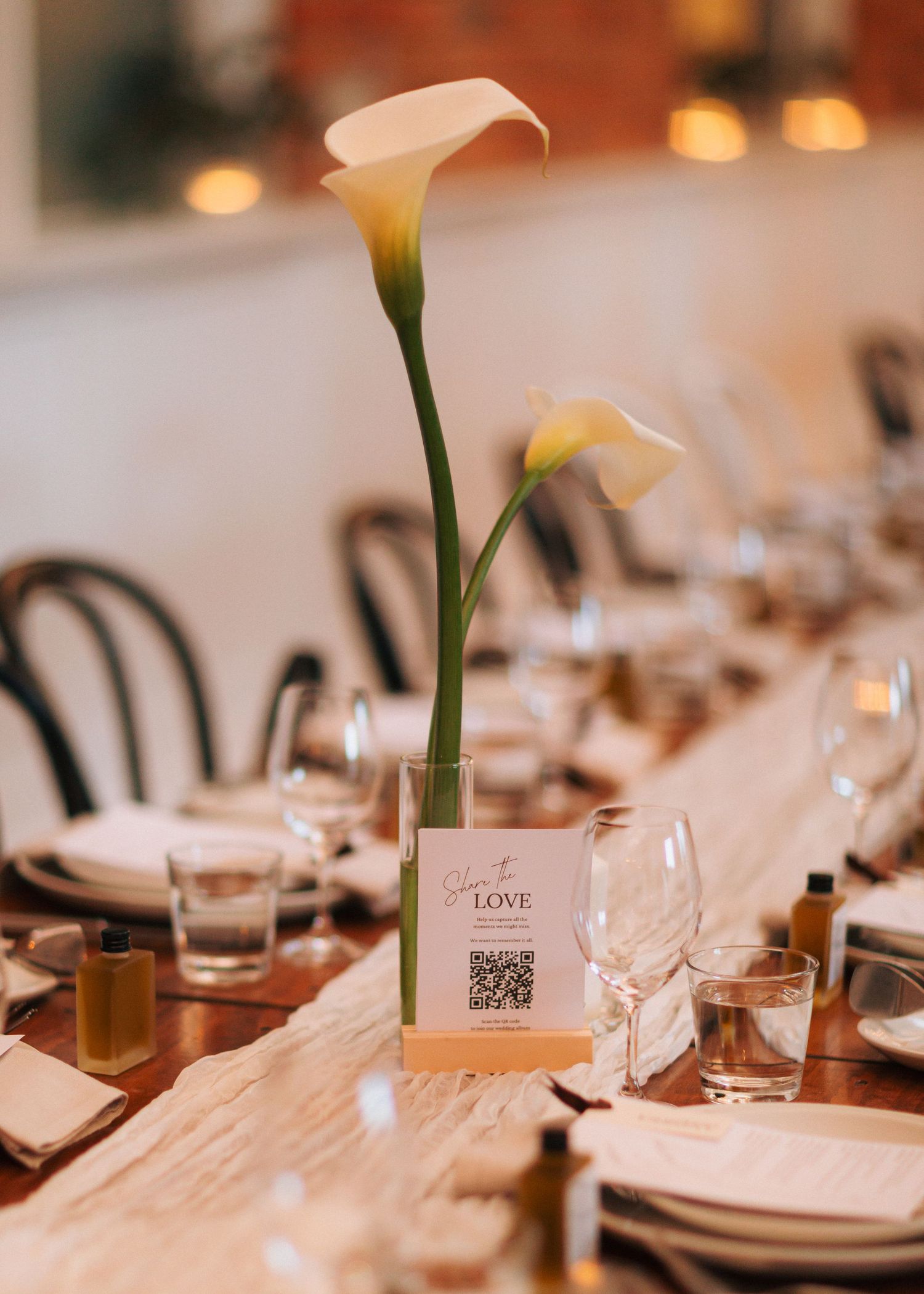 Elegant wedding reception table with calla lily centerpiece, QR code sign, wine glasses, and warm candlelight.