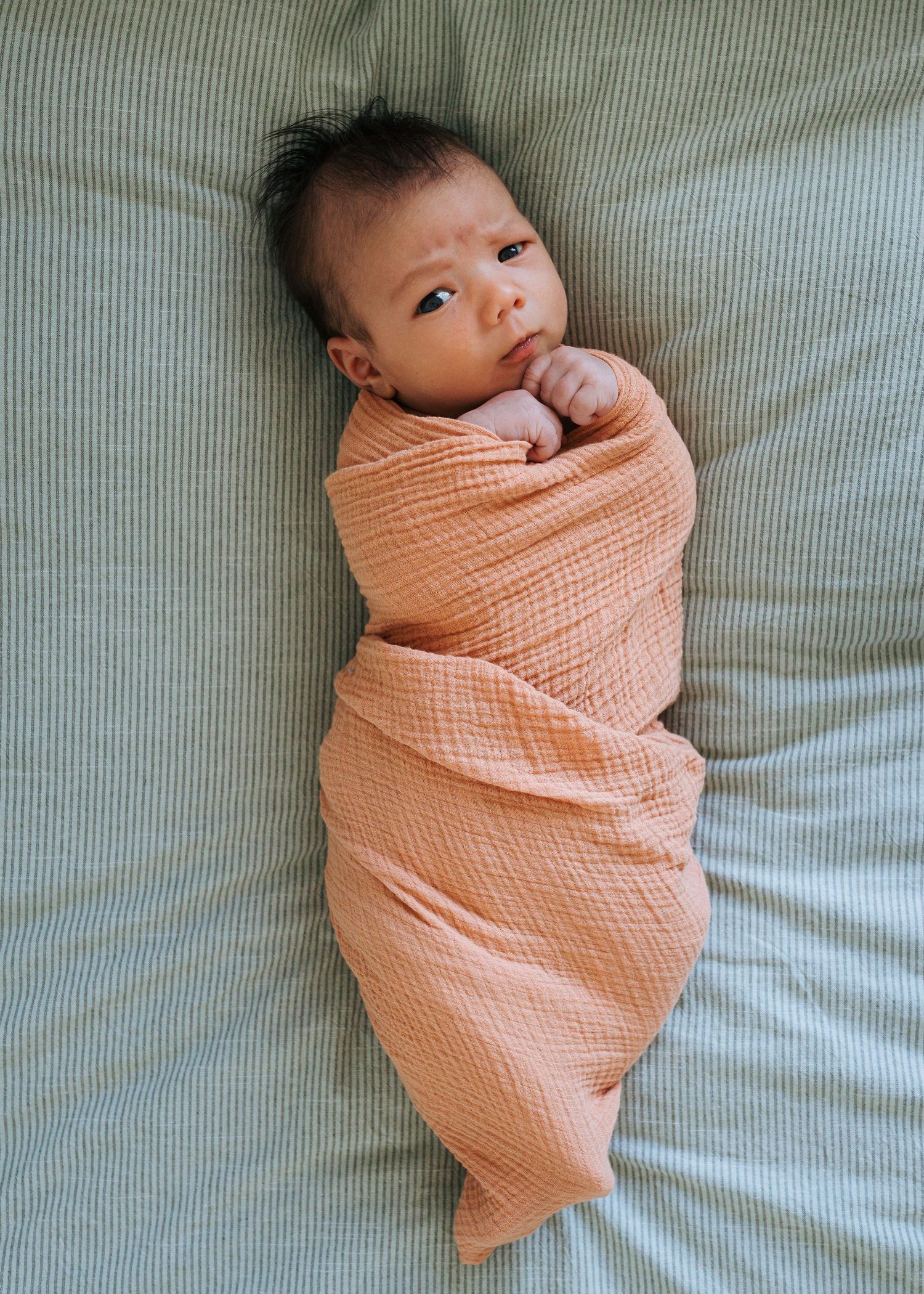 Wide-eyed newborn baby swaddled in a peach muslin blanket lies on a sage green striped sheet with hands tucked near the chin.