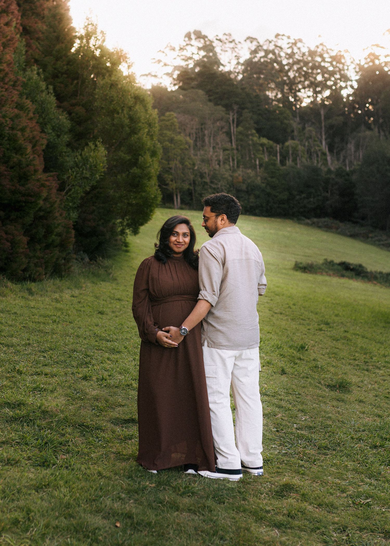 Expecting couple in a brown maxi dress and cream outfit embrace on a grassy hillside surrounded by tall green trees at sunset.
