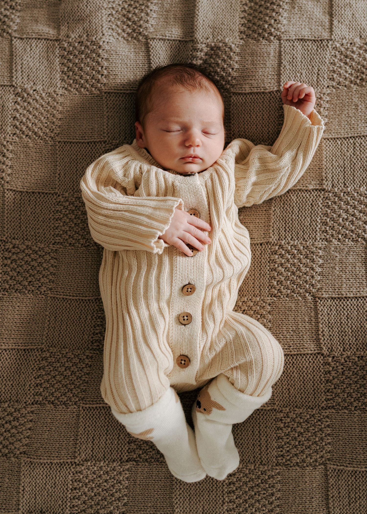 Newborn baby in cream ribbed knit romper lying on textured brown blanket with arms raised.