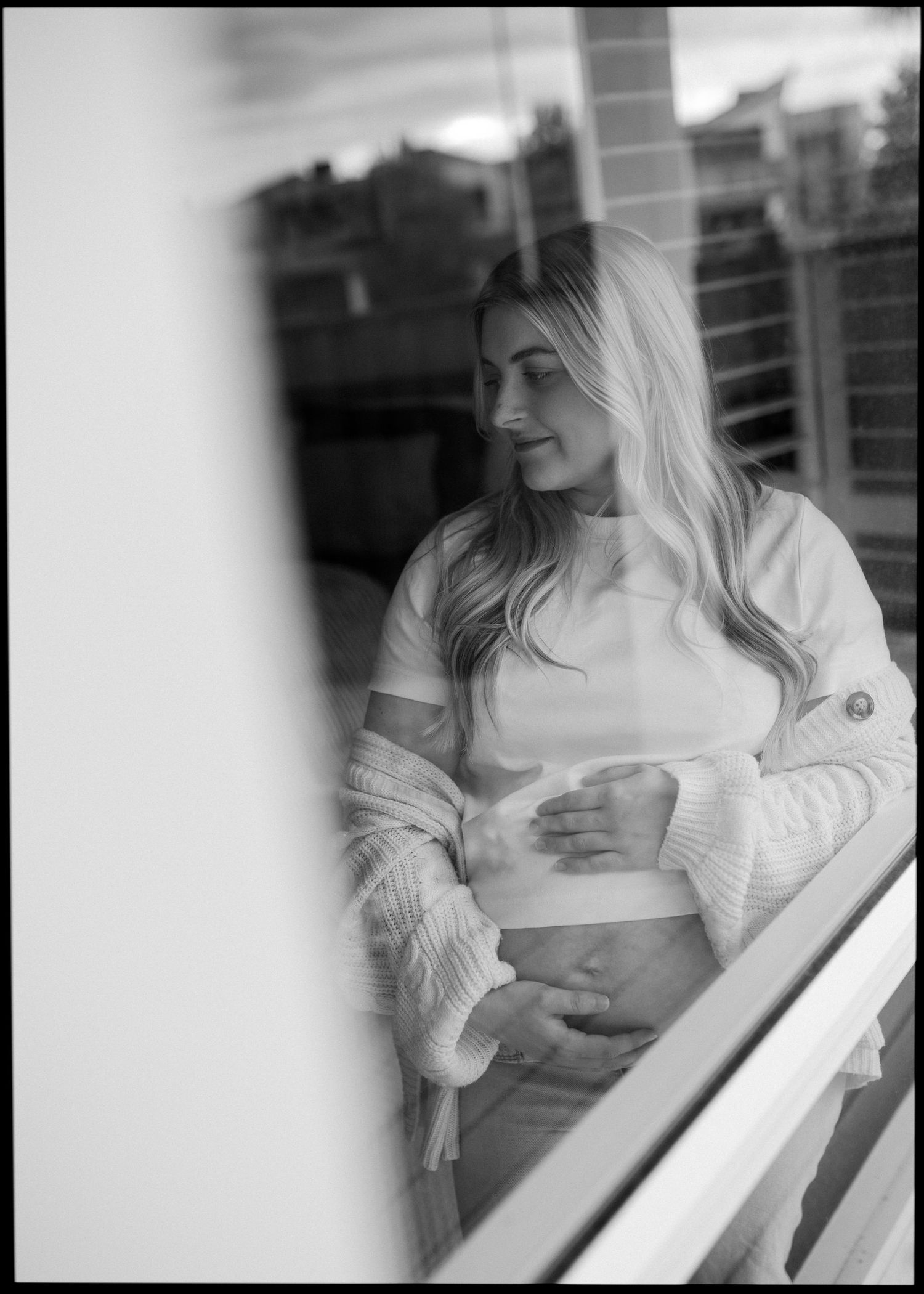 Black and white maternity photo of pregnant woman cradling bump, shot through glass with reflection.