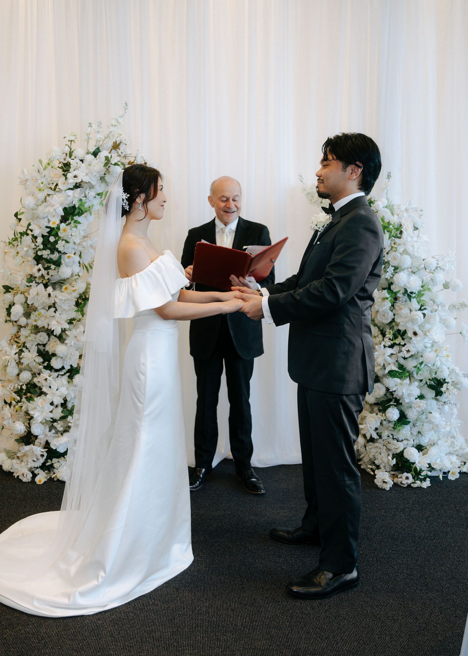 Bride and groom holding hands exchanging vows before officiant at white floral wedding ceremony altar.