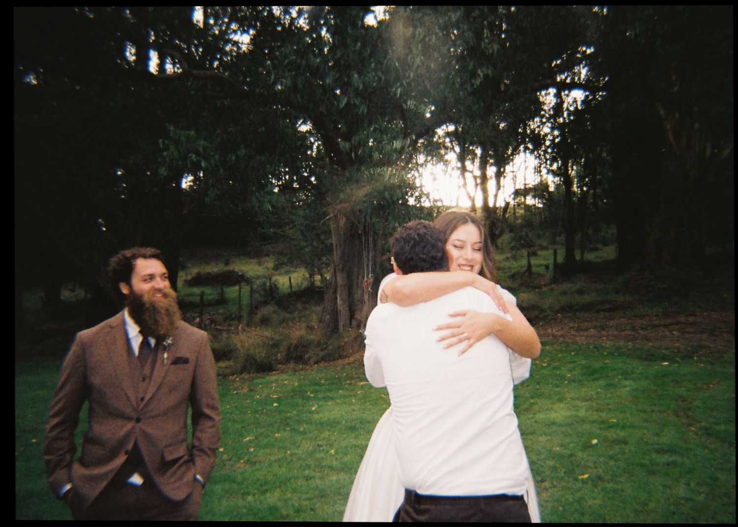 Bride in white gown sharing a warm embrace with a guest while groom in brown suit watches at sunset.