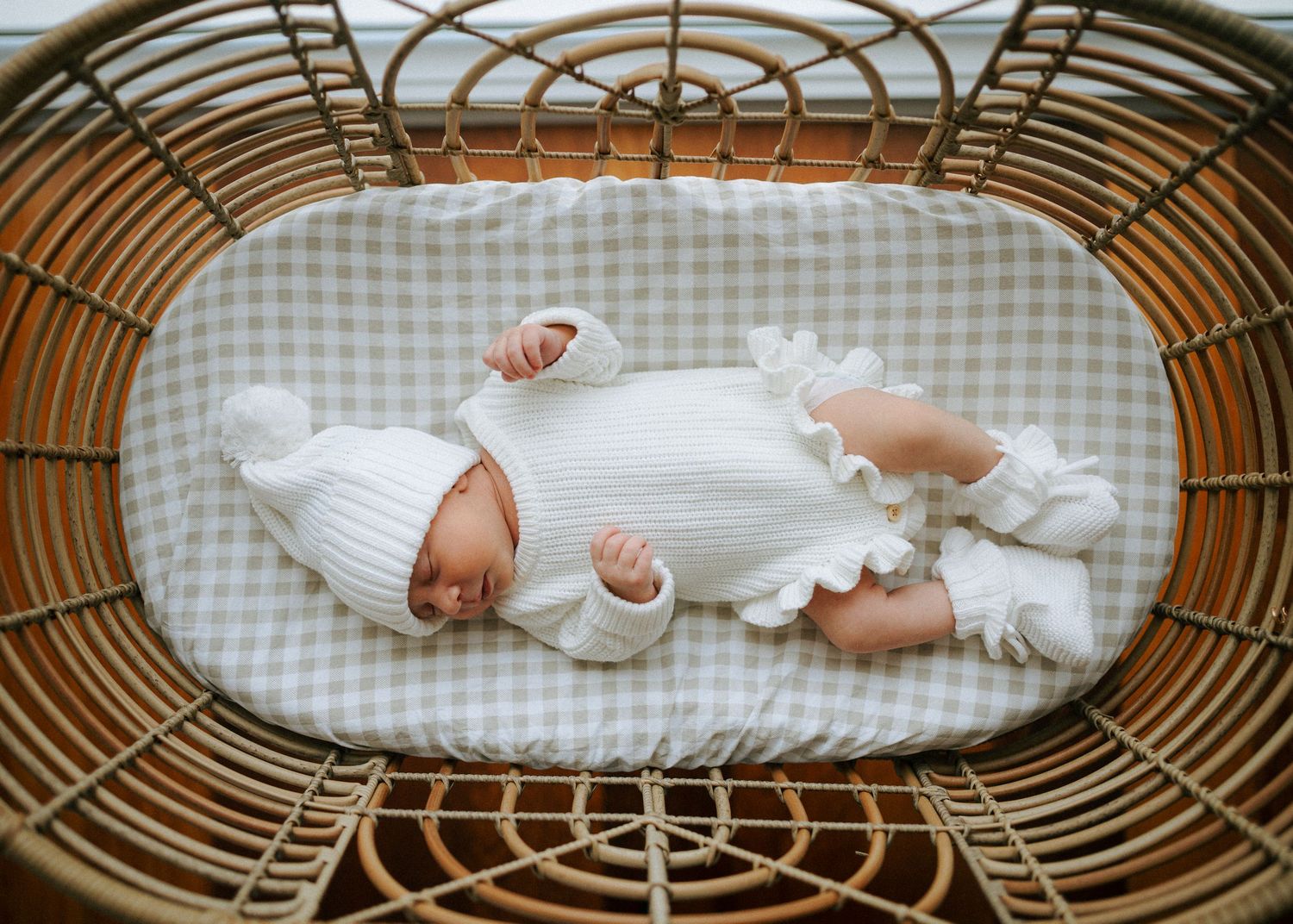 Sleeping newborn baby in white knit outfit and hat lying in a wicker bassinet on gingham bedding.
