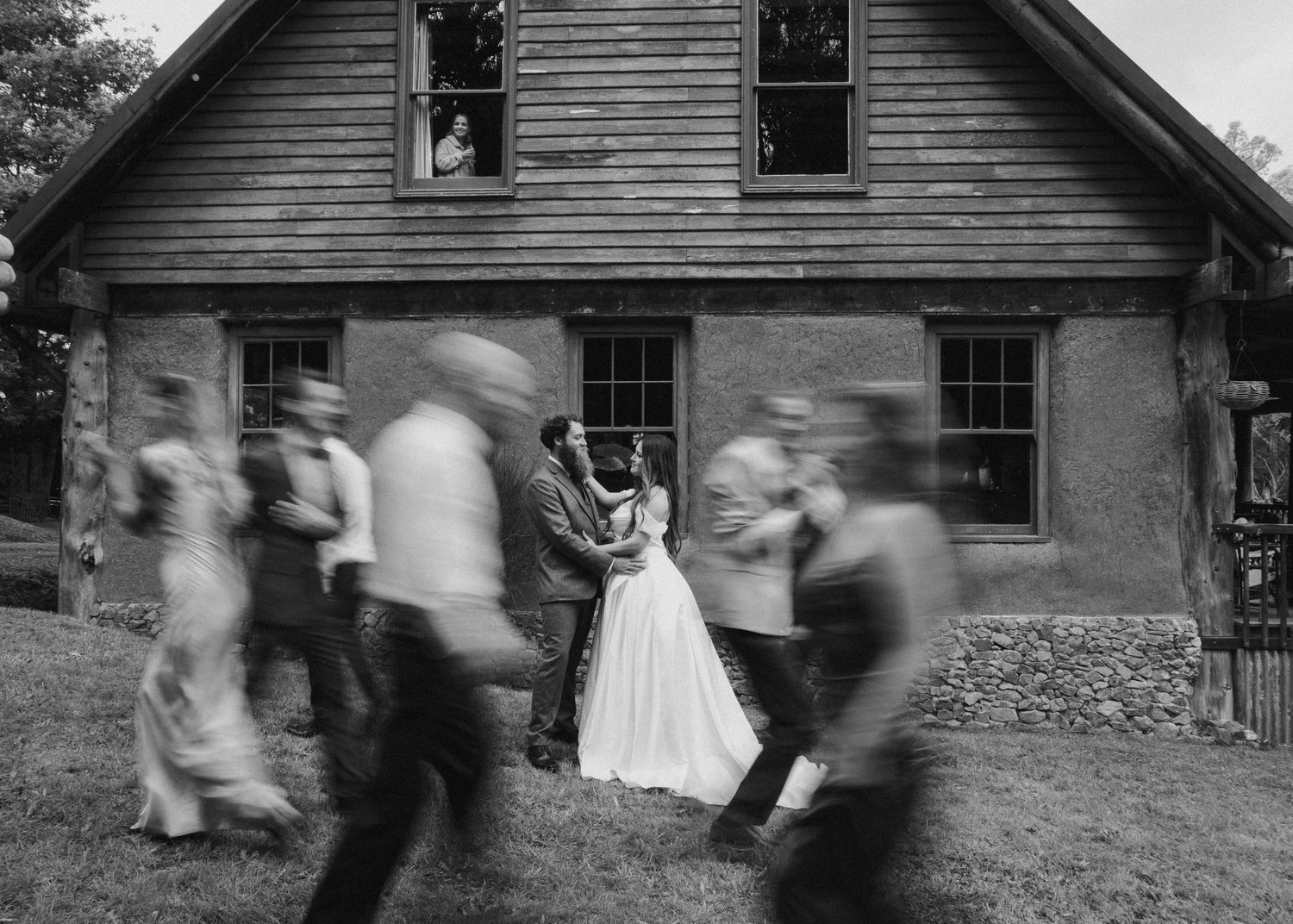 Black and white photo of bride and groom standing still as wedding party moves past in blur.