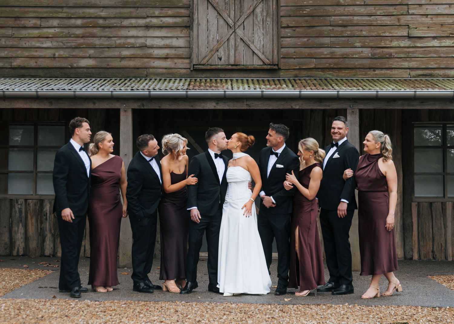 Wedding party kissing and celebrating outside rustic barn venue.