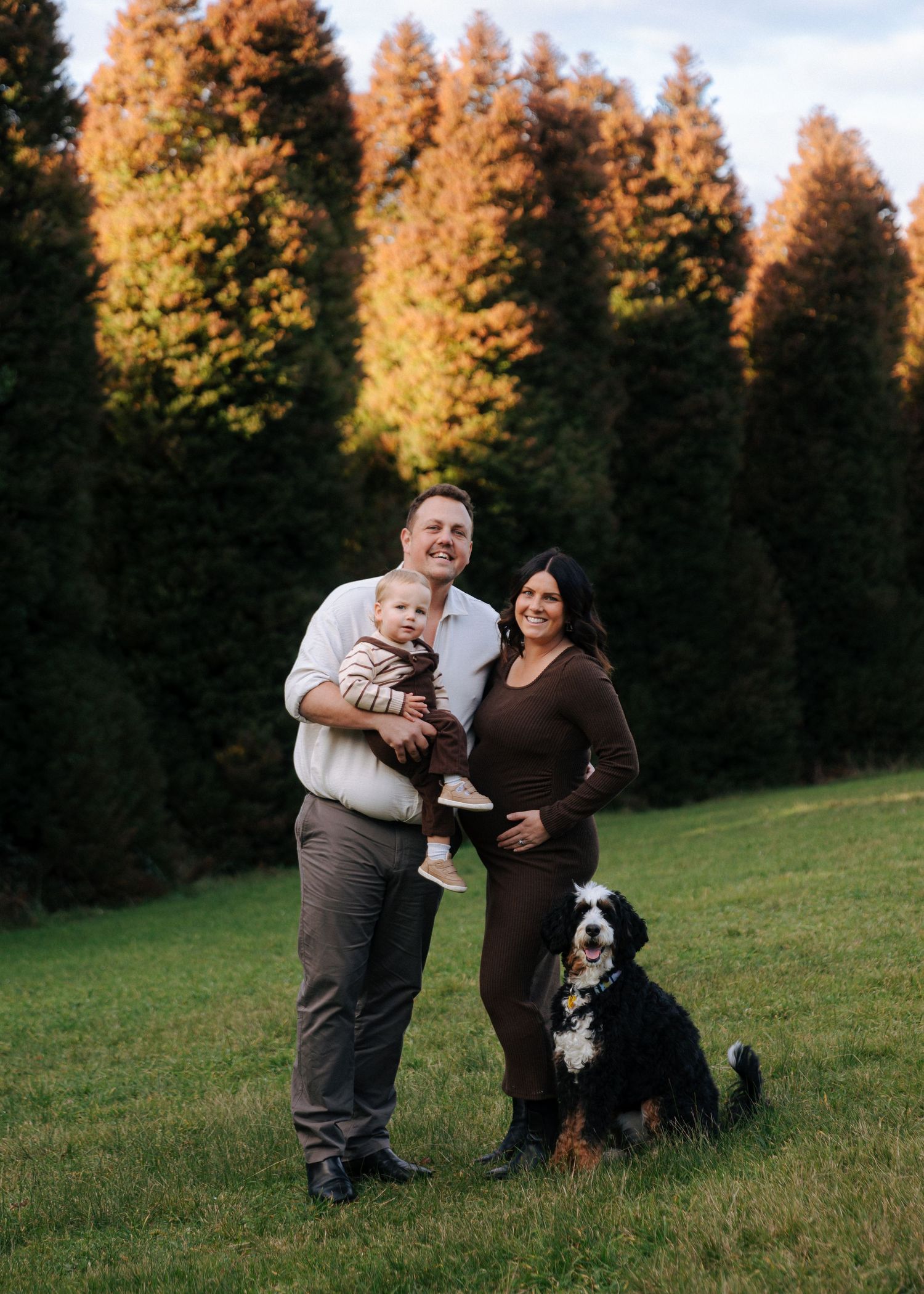 Family portrait with man, pregnant woman, baby, and Bernese Mountain Dog outdoors.