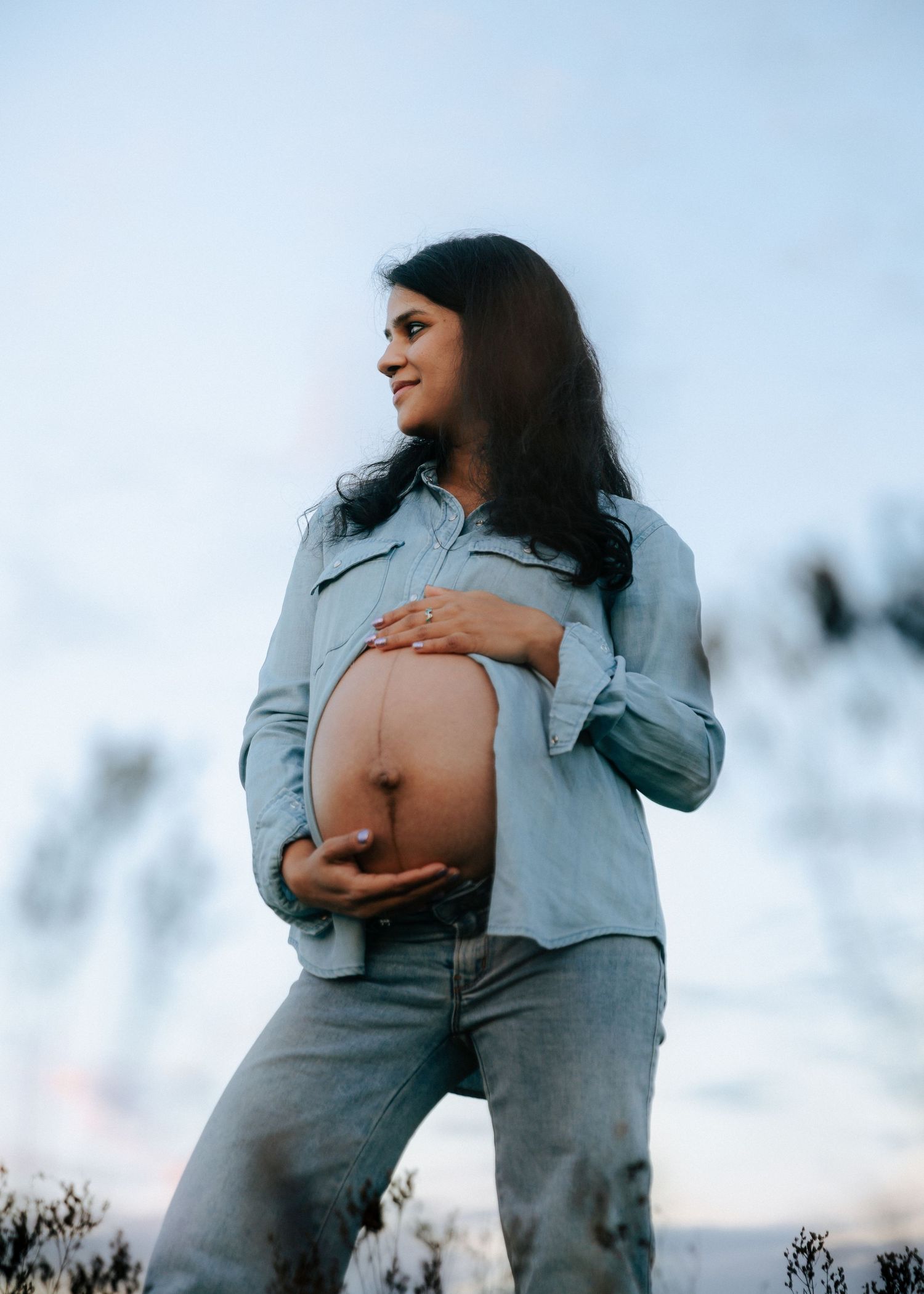 Radiant pregnant woman in light blue denim shirt cradling her bare belly against a soft sky.