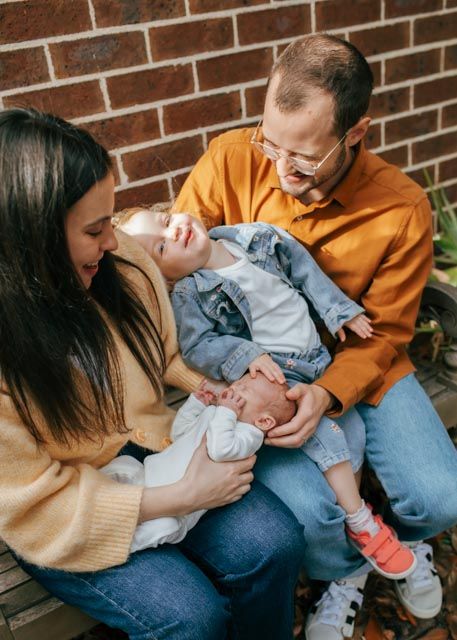 Happy family with toddler and newborn baby sitting against brick wall outdoors.