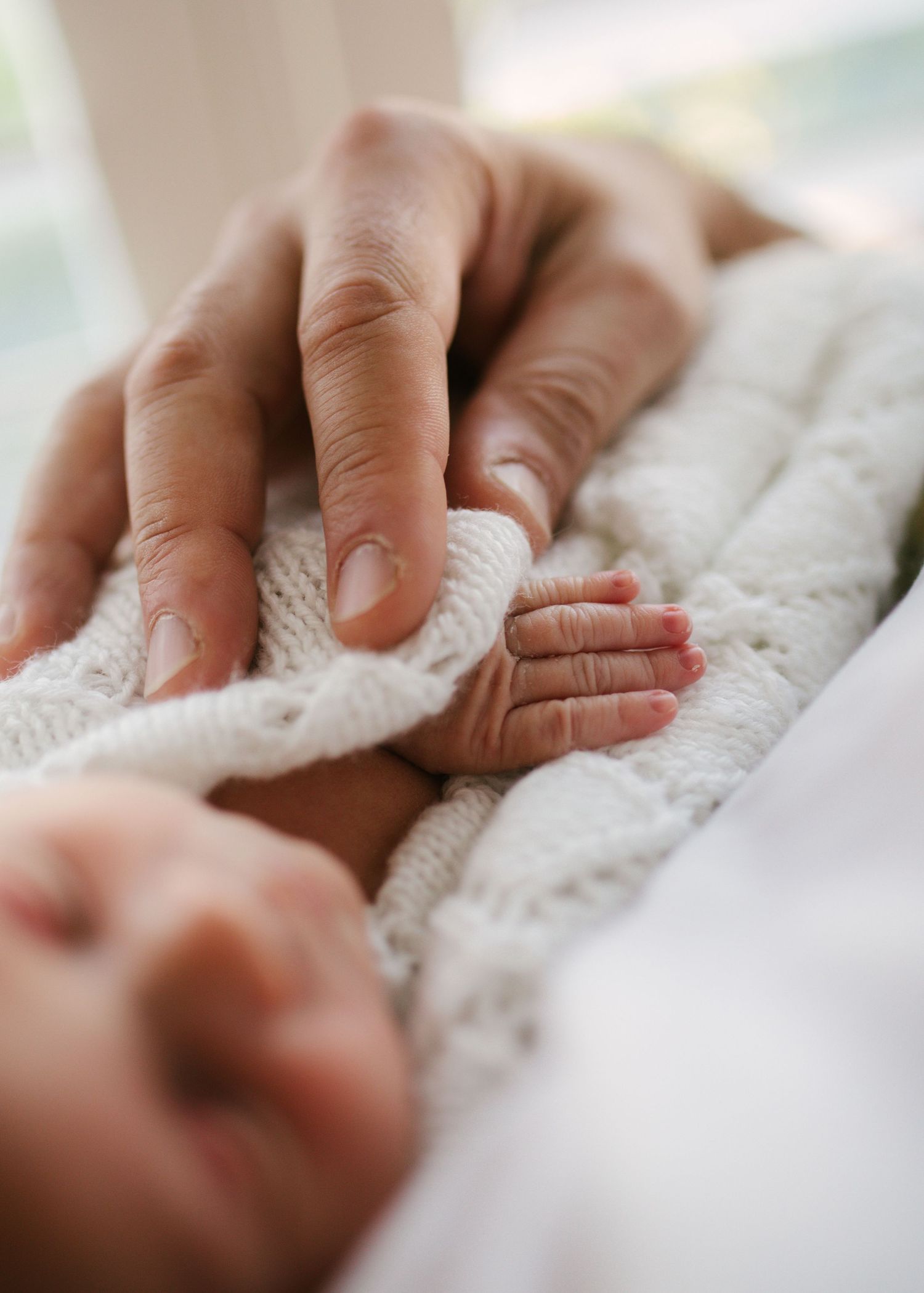 Close-up of adult hand tenderly touching tiny newborn baby foot on white knit blanket.