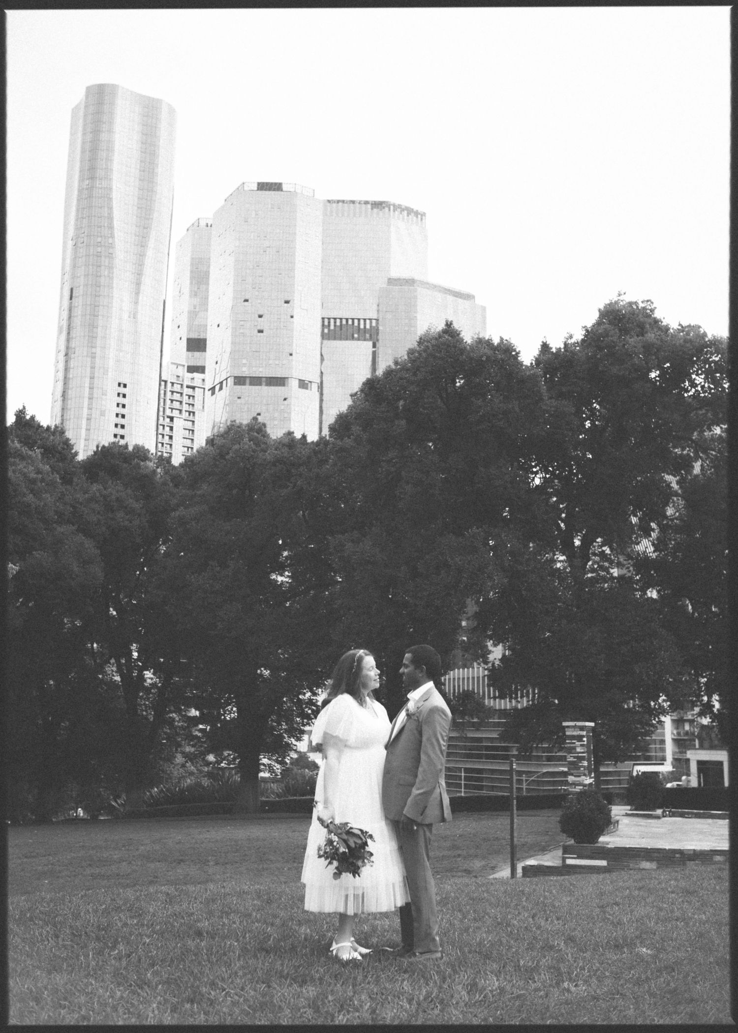 Bride and groom standing on grass with city skyscrapers in the background, black and white.