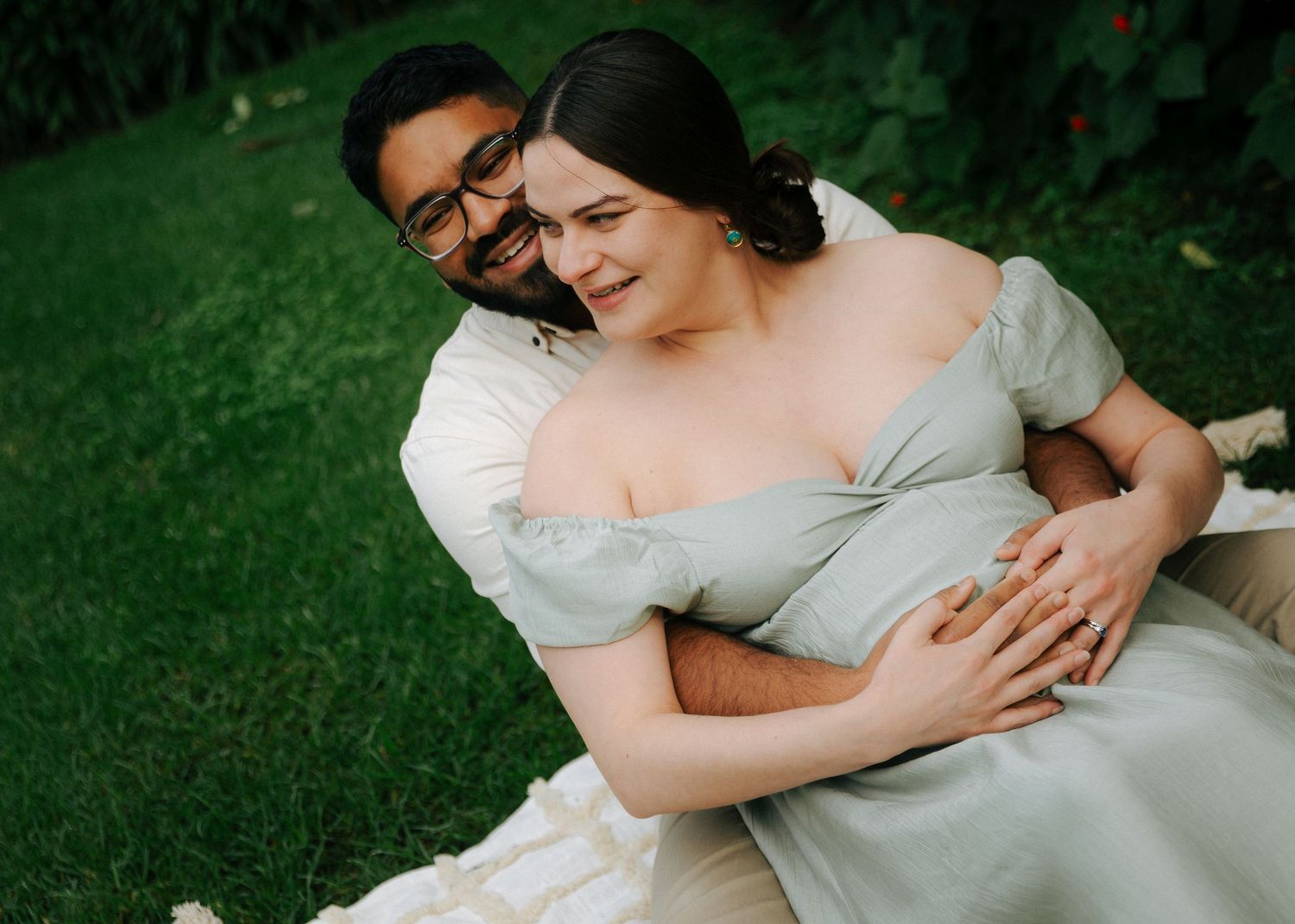 Couple sitting on grass, man embracing pregnant woman in sage green maternity dress from behind.